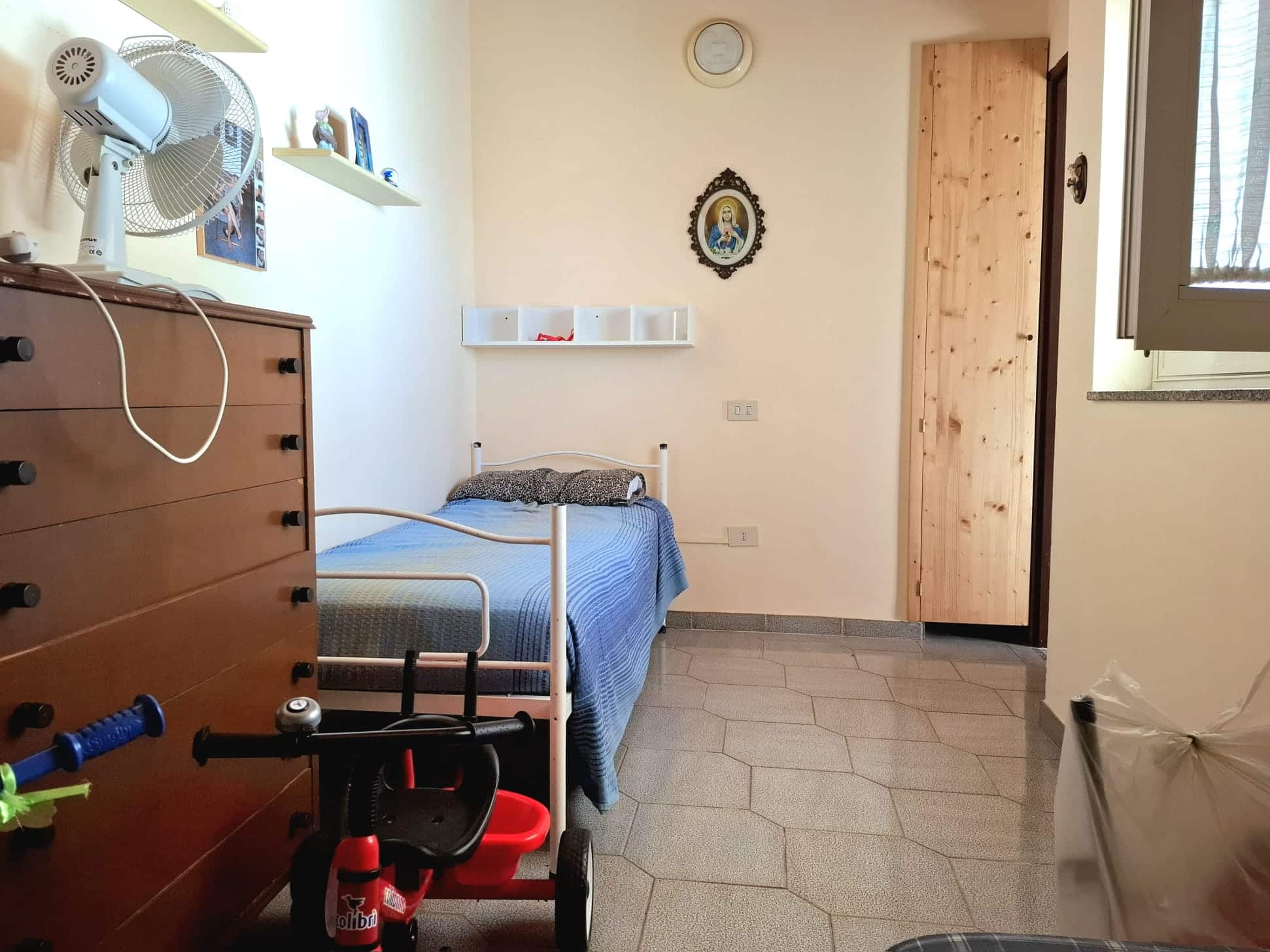 Letto singolo in camera da letto con arredi pratici e decorazioni semplici, finestra per luce naturale, mobili in legno, atmosfera accogliente e funzionale.