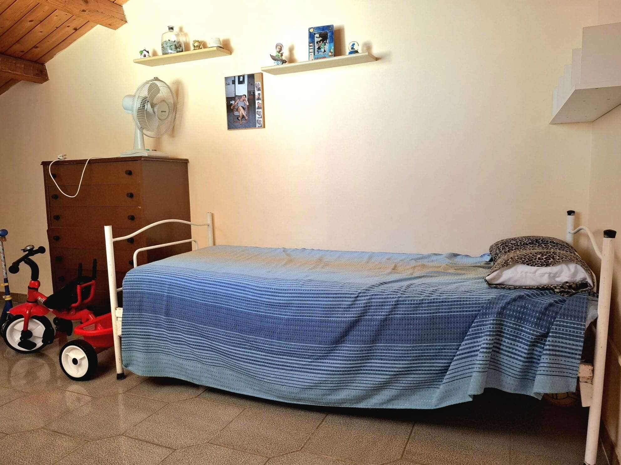 Letto singolo con copriletto blu, stanza arredata con armadio antico, ventilatore e decorazioni wall art, spazio dedicato alla casa in stile funzionale e accogliente.
