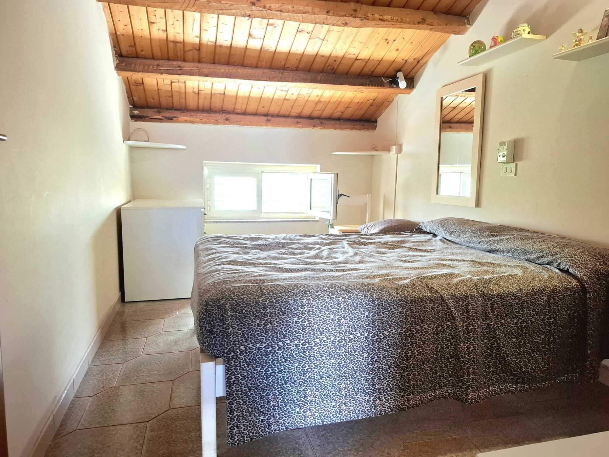 Camera da letto con soffitto a travi in legno, letto matrimoniale, pareti chiare e finestra che permette l'ingresso di luce naturale, arredi semplici e funzionali, atmosfera accogliente e luminosa.