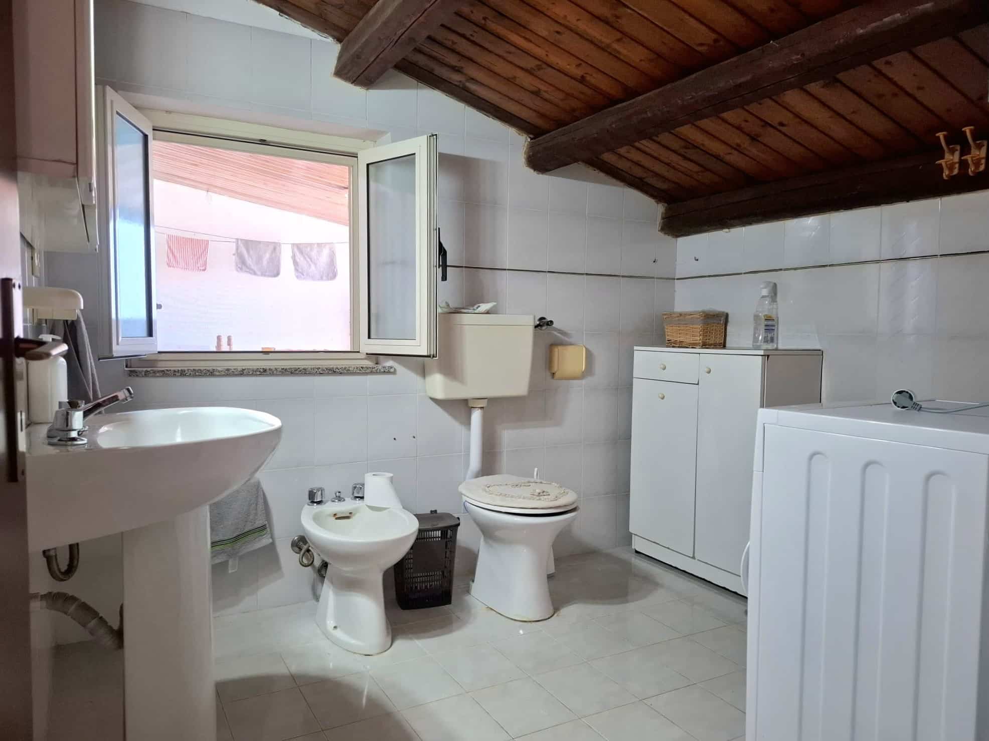 Vecchia stanza da bagno con sanitari bianchi, finestra aperta, soffitto in legno e pareti piastrellate, ideale per ristrutturazione o esempio di design classico.