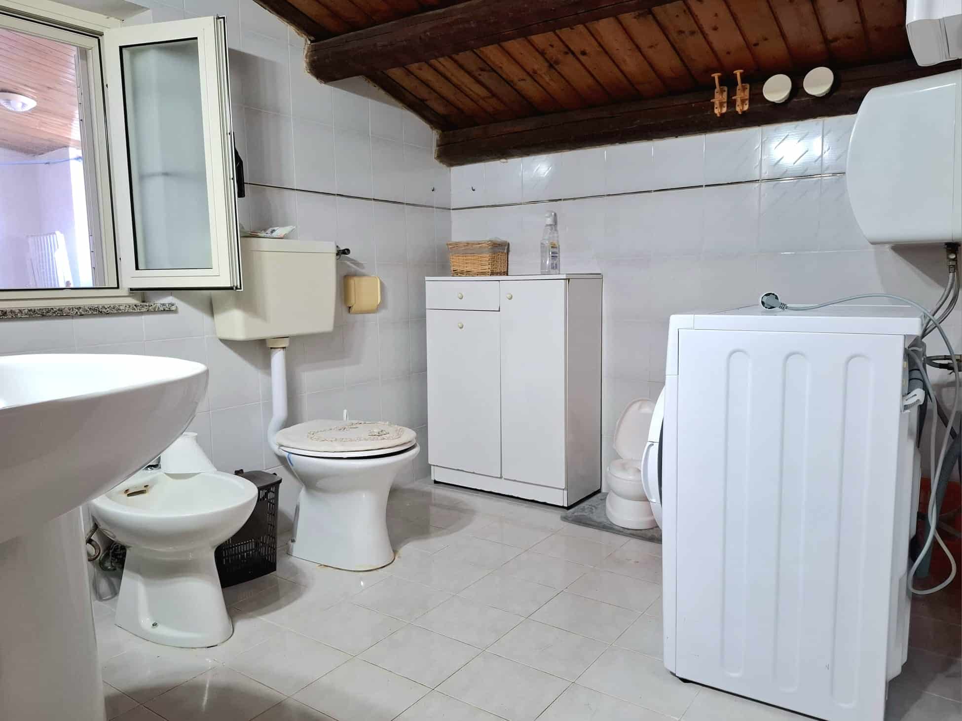 Lavandino, toilette e lavatrice in bagno con pareti rivestite di piastrelle bianche e soffitto in legno, ambiente funzionale e pratico per le esigenze quotidiane.