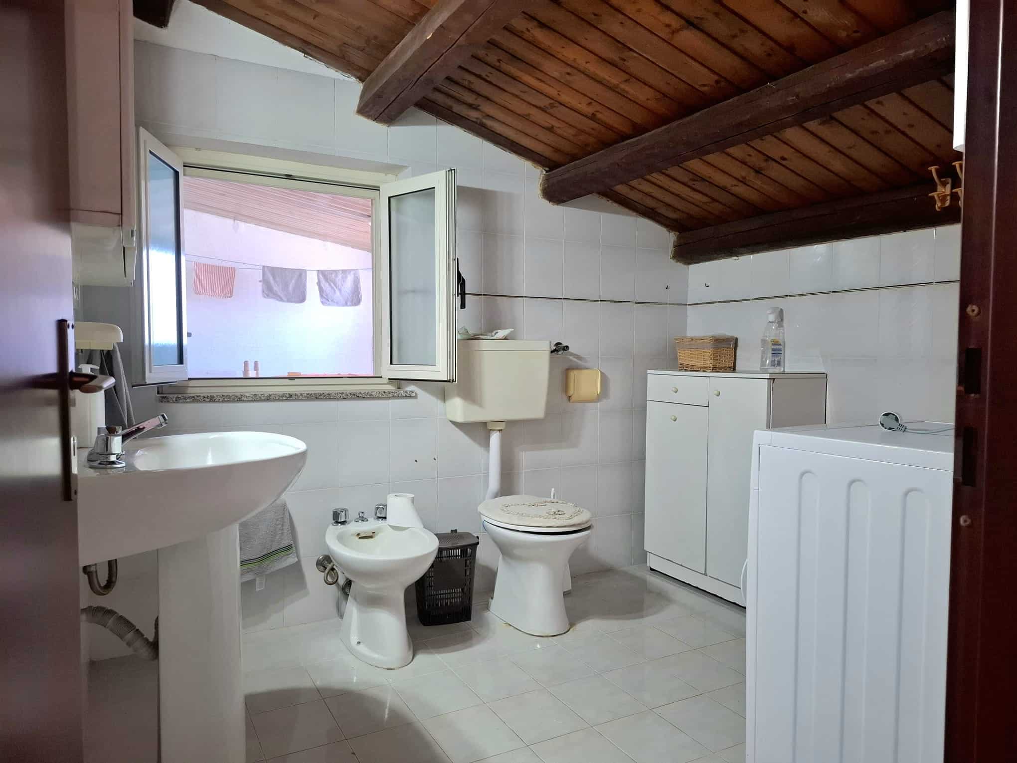Lavabo, bidet, toilette e lavatrice in bagno con soffitto in legno e finestra apribile, ambienti luminosi e funzionali, ideale per ristrutturazione o seconda casa.
