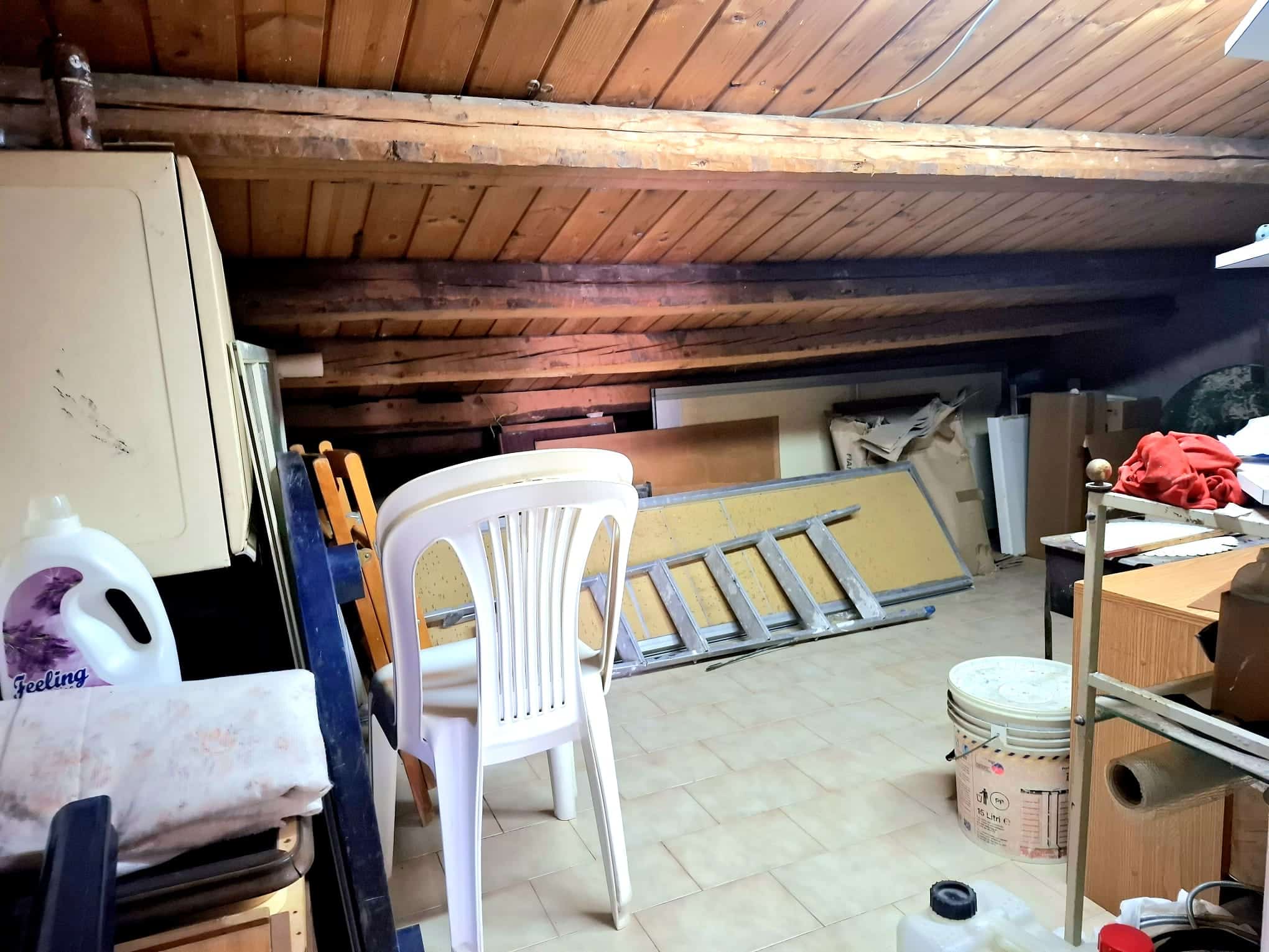 L'immagine mostra un garage o magazzino con soffitto in legno, contenente materiali di bricolage, mobili e vari strumenti di deposito, creando uno spazio funzionale per il stoccaggio e il fai-da-te.