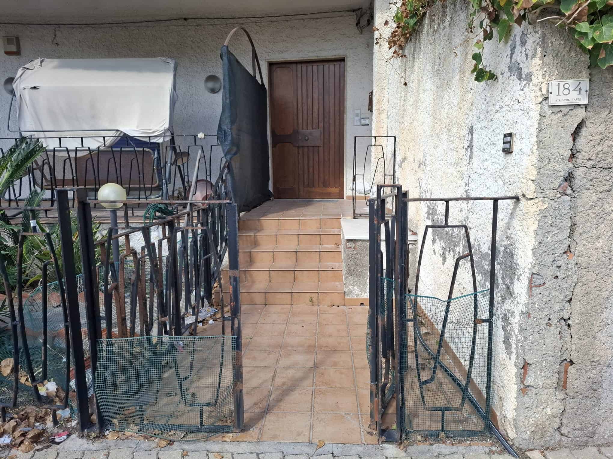 Porticato esterno di casa con ringhiera in ferro, scala in piastrelle, porta in legno e pareti in cemento, situato in zona urbana con dettagli di foglie e elementi di arredo.