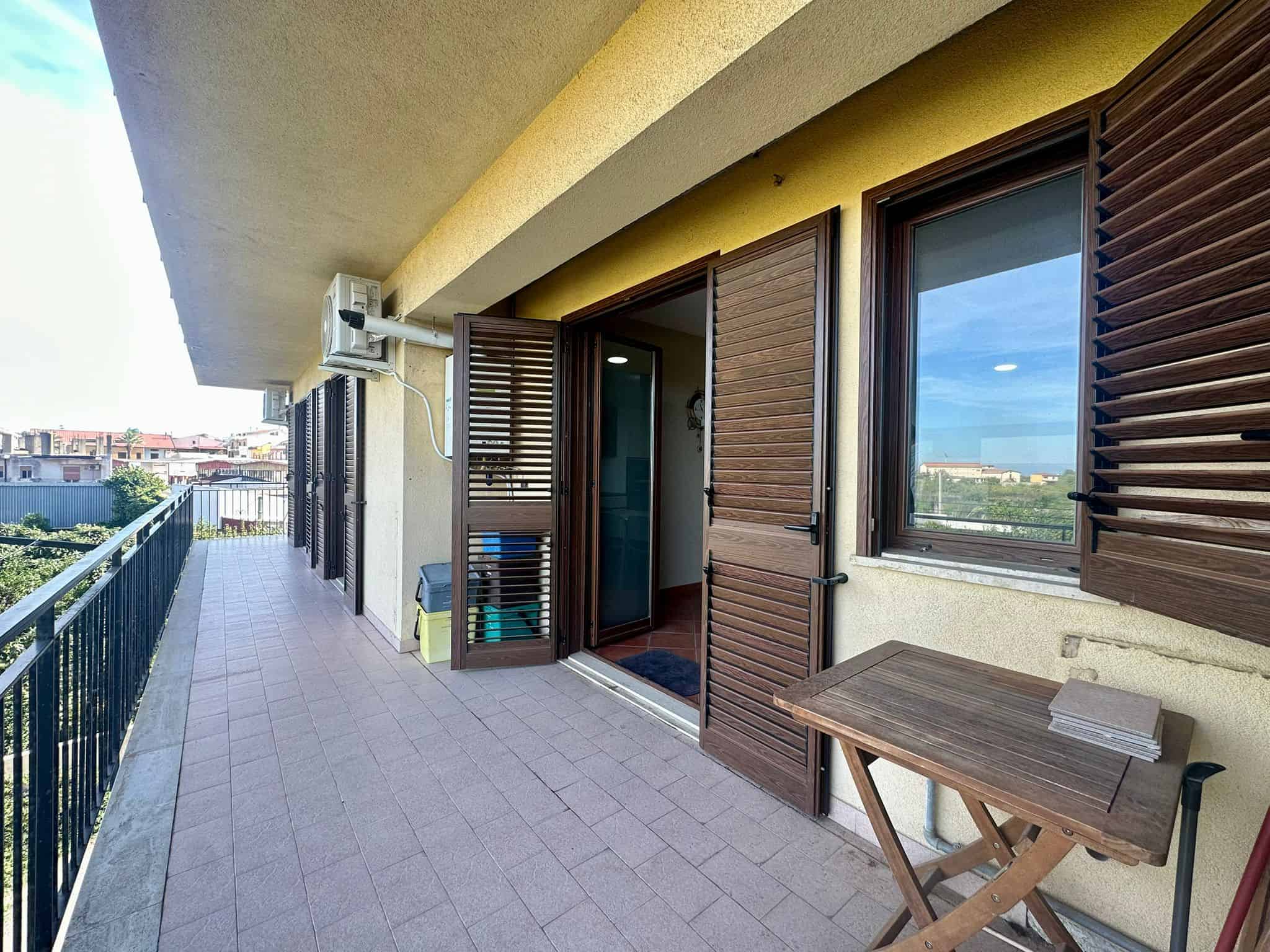 Balcone spazioso con finestre a vetro e persiane in legno, ideale per relax e momenti all'aperto, vista su zona urbana e verde, accesso diretto dalla casa, spazio per mobili e piante.