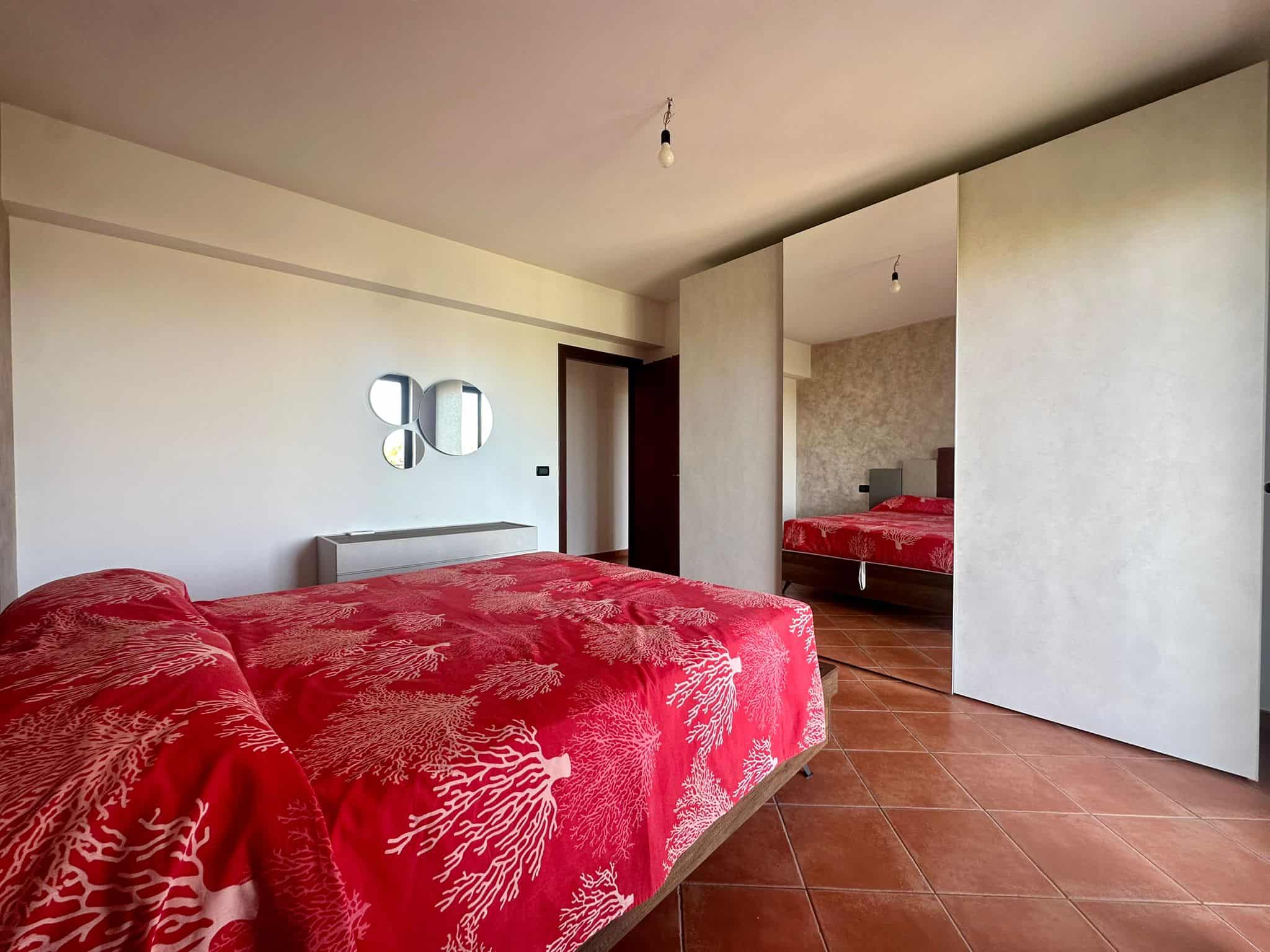 Letto matrimoniale con coperta rossa a motivi coraline, camera spaziosa con pareti chiare e specchio decorativo, arredi moderni, parquet chiaro, ambiente luminoso e accogliente.