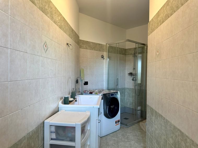 Lavanderia con box doccia e lavatrice, pavimenti in ceramica e pareti piastrellate, spazio funzionale e ben illuminato, ideale per l'organizzazione domestica quotidiana.