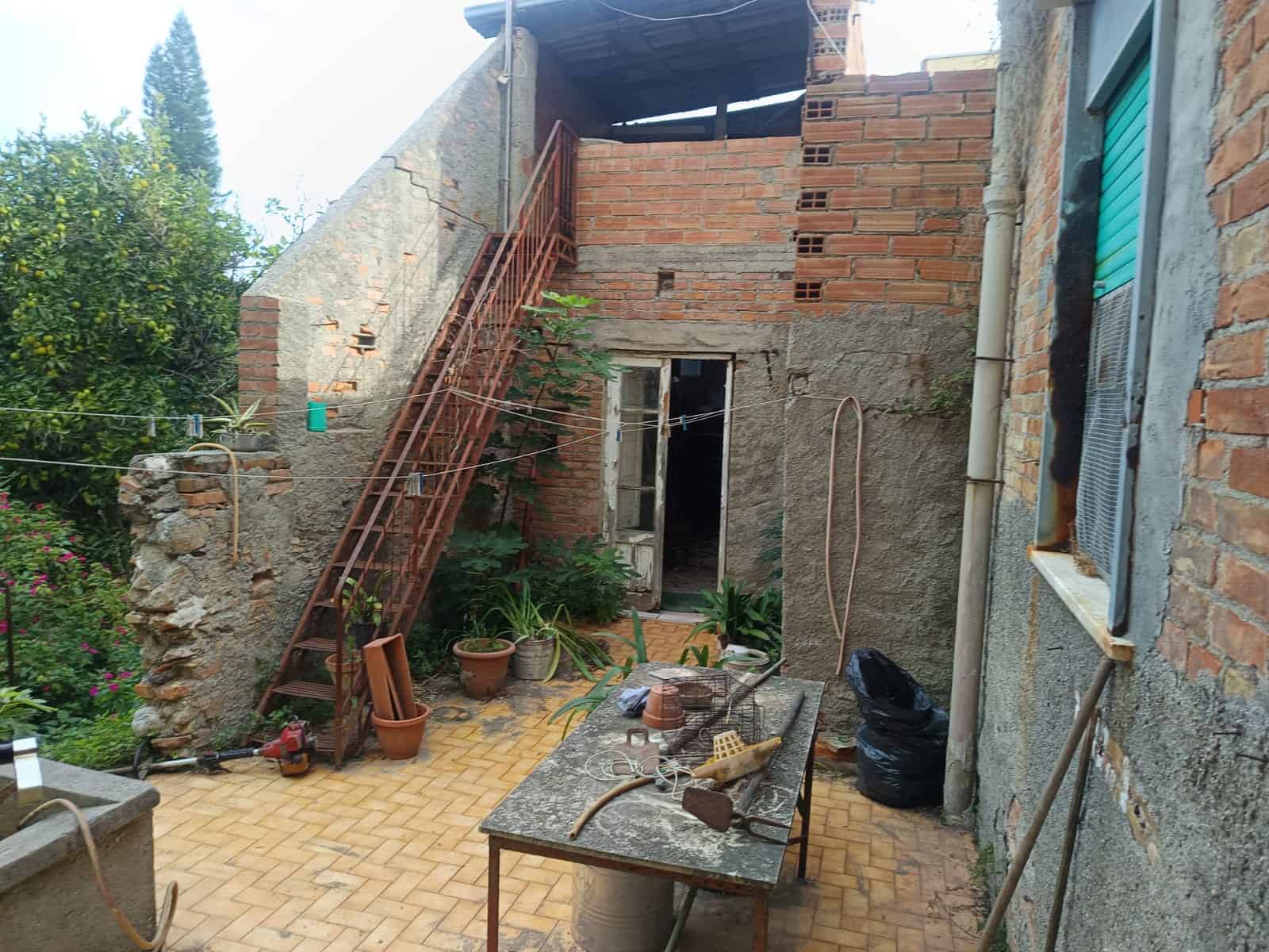 Scala di ferro esterna di una casa in fase di ristrutturazione, con assi di legno, ringhiera rossa e piante in vaso, su un cortile con mattonelle gialle; lavori di edilizia e ristrutturazione.