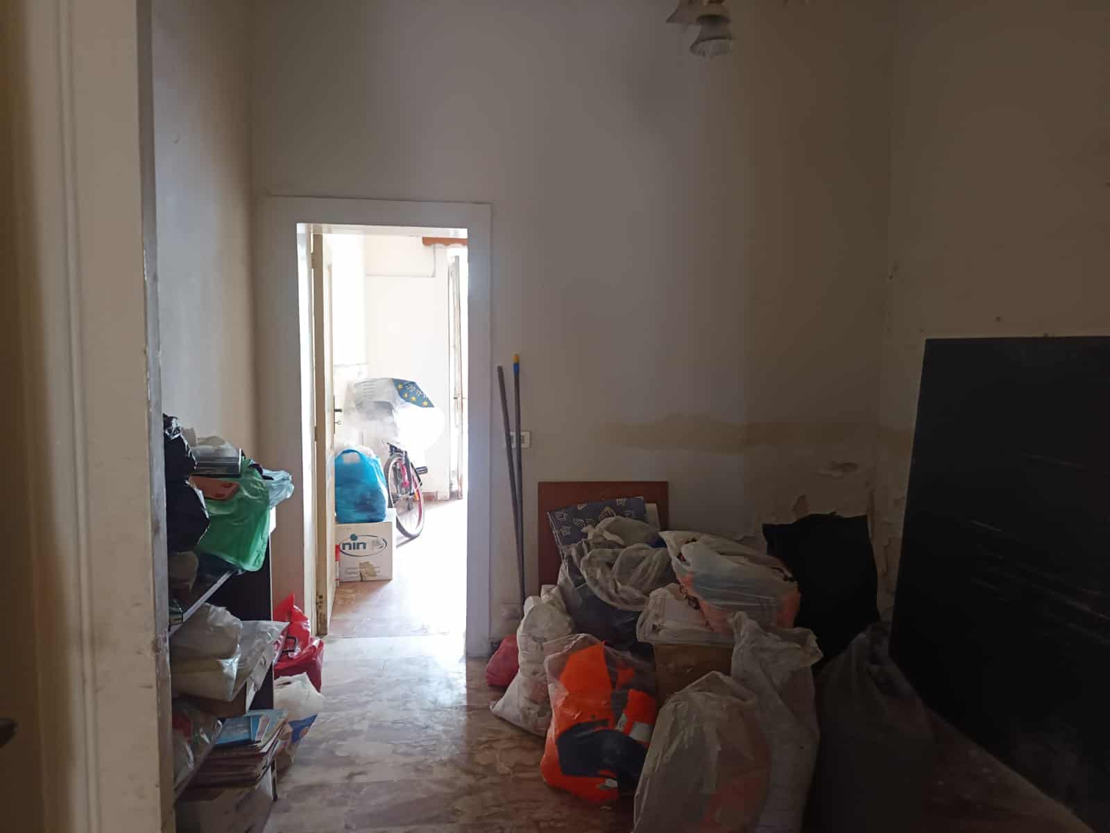Punto vendita NextCasa, immobile da ristrutturare con spazio di deposito, stanza disordinata con sacchi della spazzatura, oggetti vari e ingresso con luce naturale, pronta per intervento di ristrutturazione.