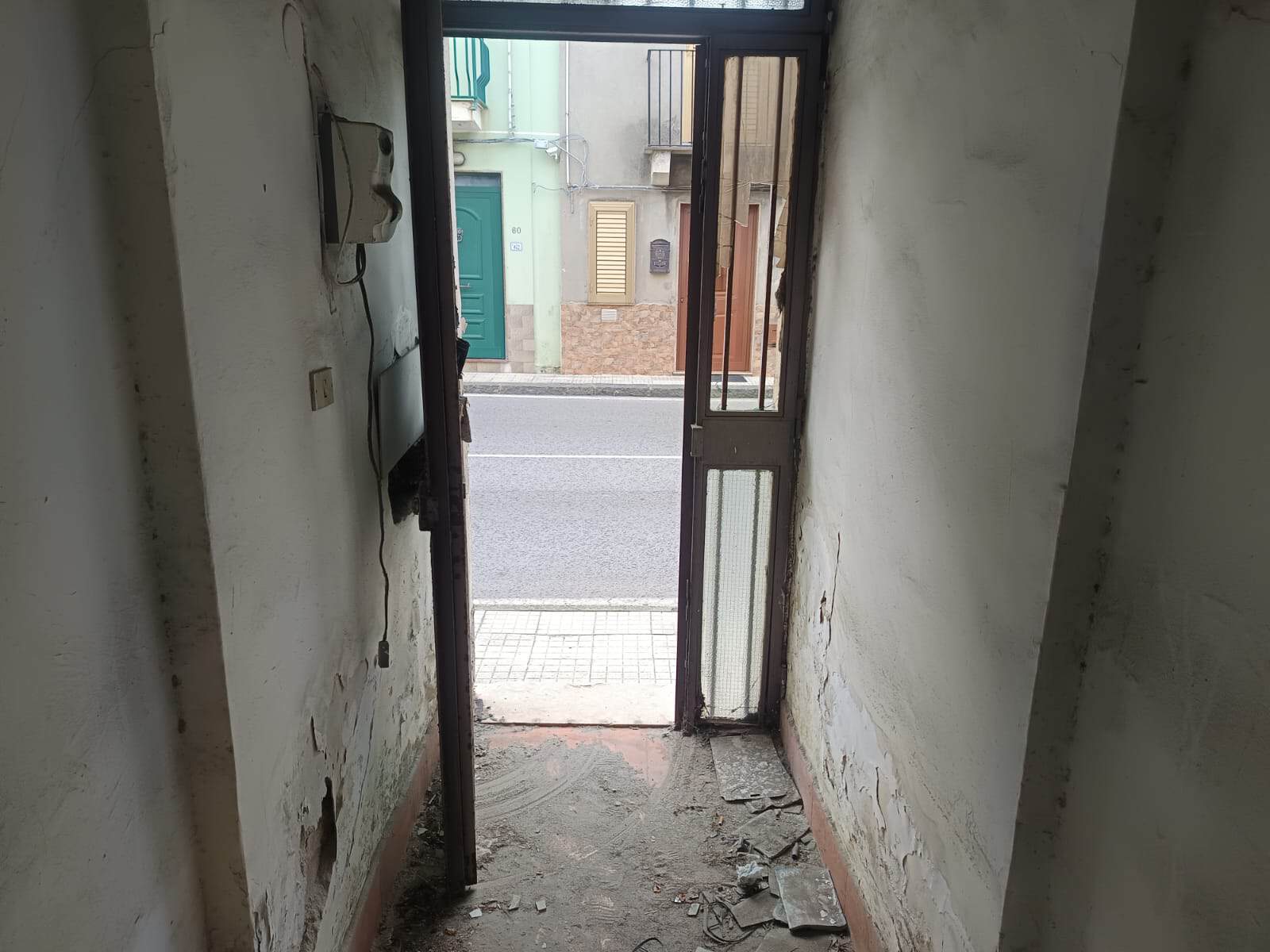 Dentro di un ingresso di una casa in ristrutturazione, pareti danneggiate e cavi elettrici scoperti, vista verso la strada con edifici colorati e porta di ingresso in legno, ambiente da rinnovare e pronto per lavori di manutenzione e ristrutturazione.