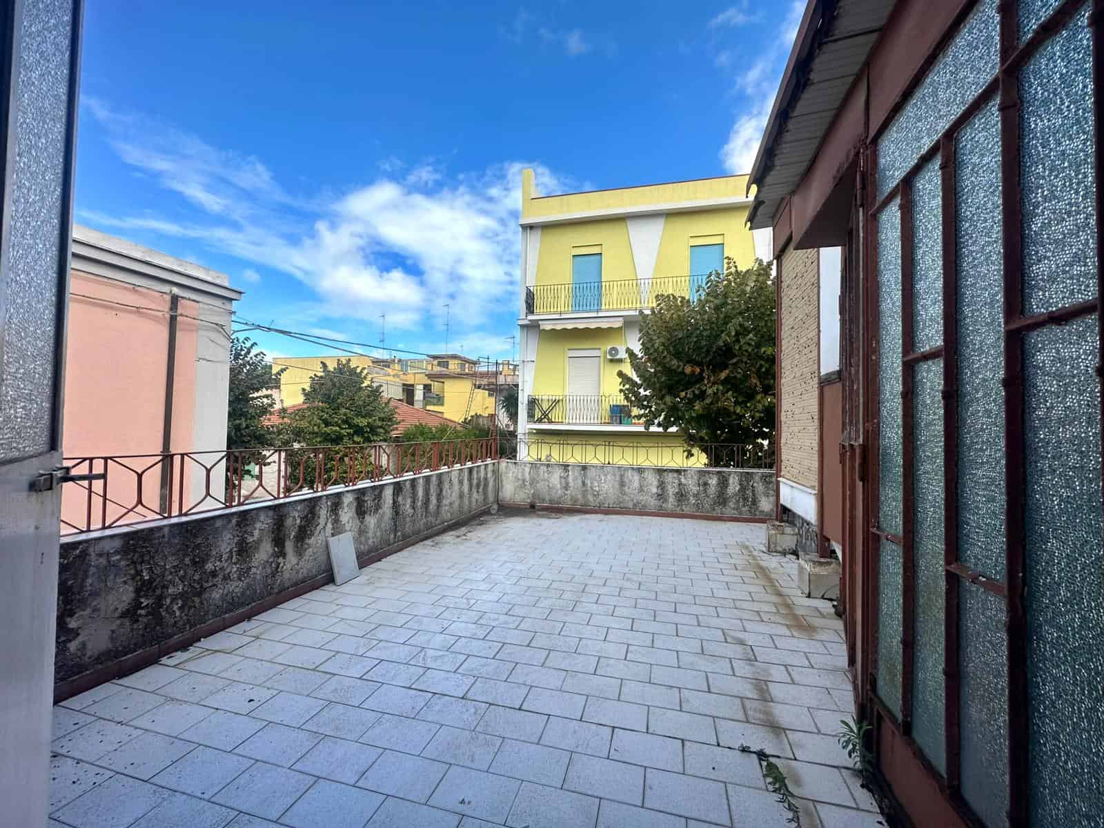 Ampio terrazzo con vista su edificio residenziale, pavimentato con piastrelle chiare, balcone e cielo sereno con nuvole sparse, perfetto per uso abitativo e relax all'aperto.