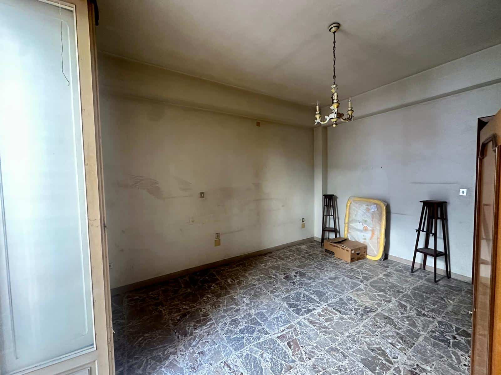 Vecchio soggiorno vuoto con pareti spente e pavimento in marmo, camera da letto da rinnovare, elementi d'epoca come lampadario e mobili vintage, spazio abitativo da modernizzare.