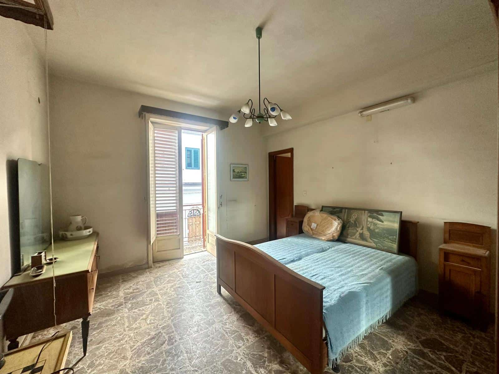 Vecchio appartamento arredato con balcone, camera da letto e mobili in legno, illuminato da luce naturale, in zona residenziale, ideale per investimento o prima casa, con ottima esposizione solare.