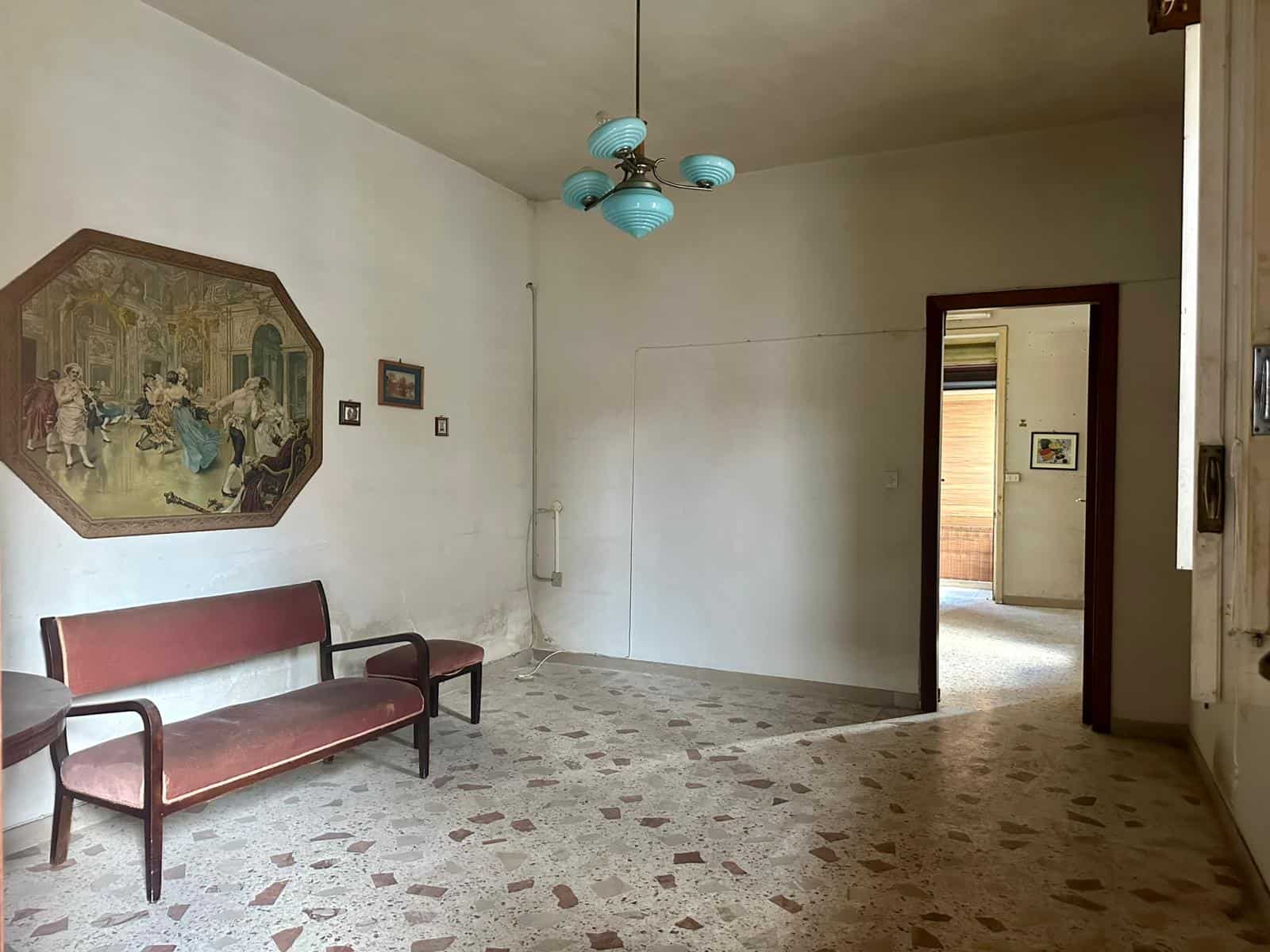 Vecchio appartamento con sala soggiorno, arredamento vintage, pareti bianche, soffitto con lampadario blu, mobili in velluto rosso, pavimento in marmo colorato, ingresso e fine stanza con vista su balcone.