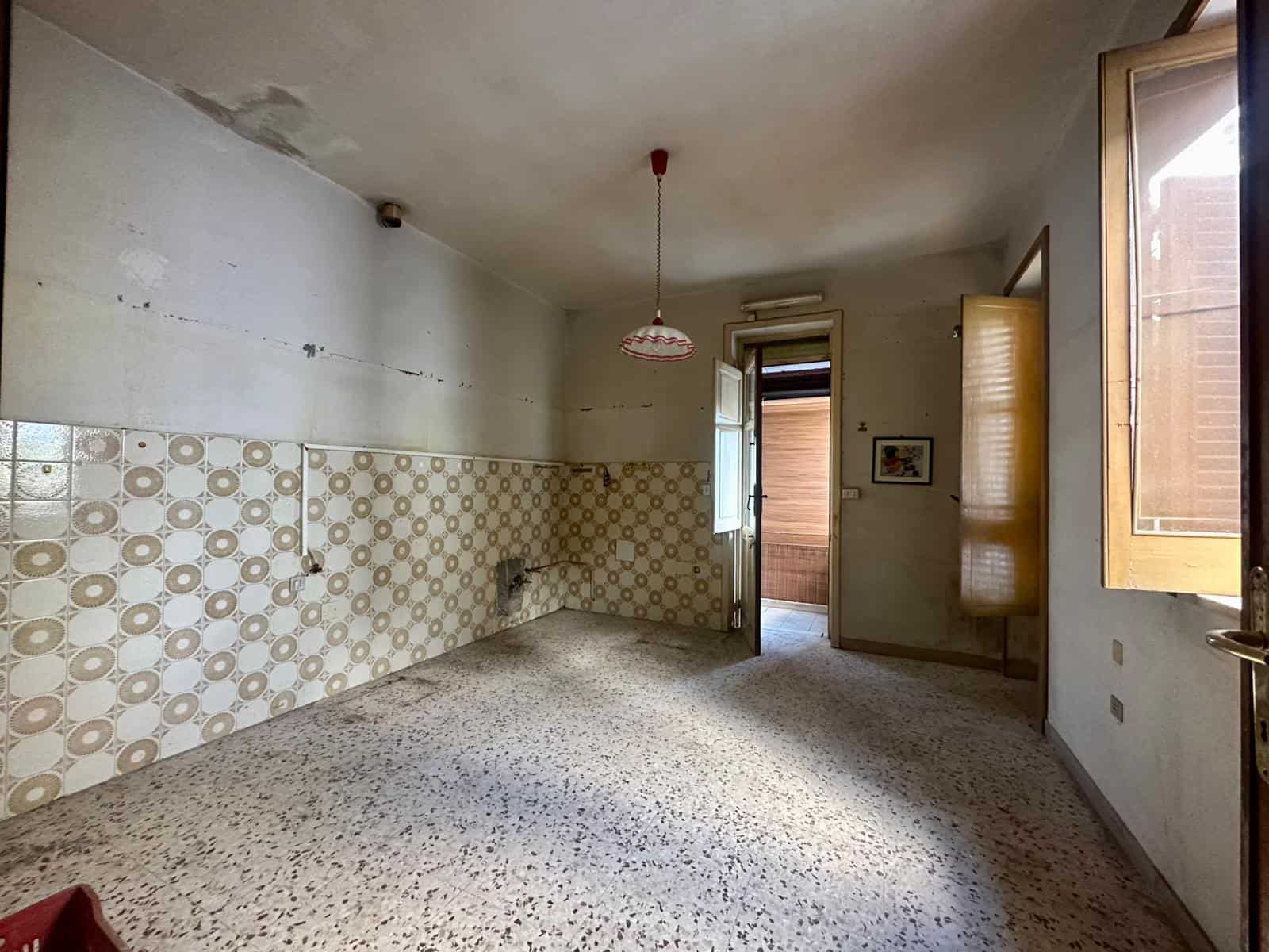 Vecchia cucina vuota in appartamento, pareti con piastrelle beige e pavimento in graniglia, finestra e porta che portano a un balcone, ambiente da ristrutturare, immobile di interesse immobiliare NextCasa.