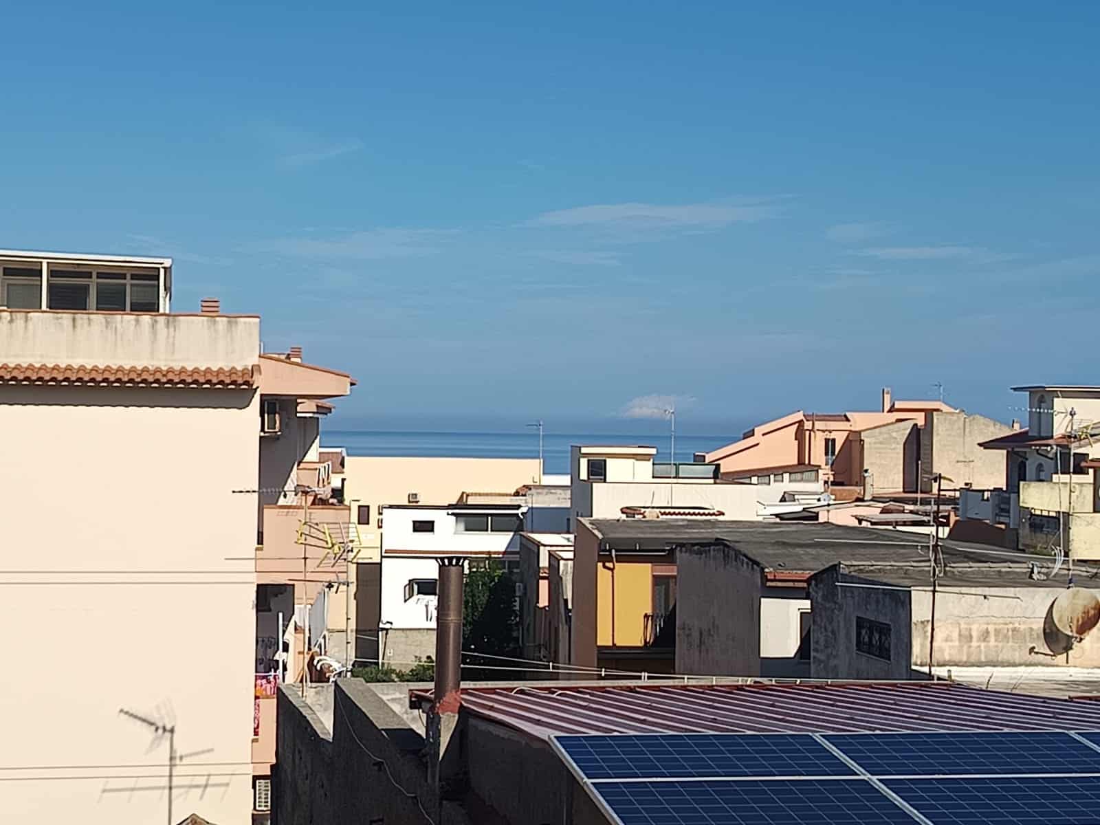 €ALT: Veduta di tetti di case residenziali con il mare sullo sfondo, cielo blu e poche nuvole, in una zona urbana soleggiata vicino alla costa.