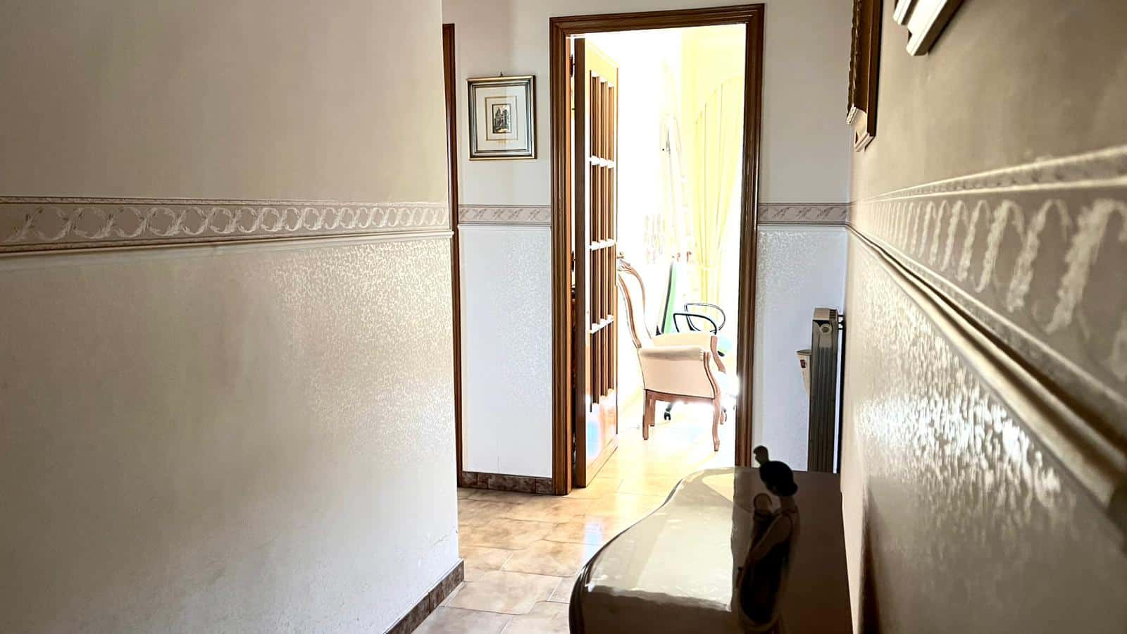 Finestra aperta su un balcone con sedie e tende gialle, illuminando un corridoio di casa arredato in stile classico con pareti decorate e oggetti d'antiquariato.