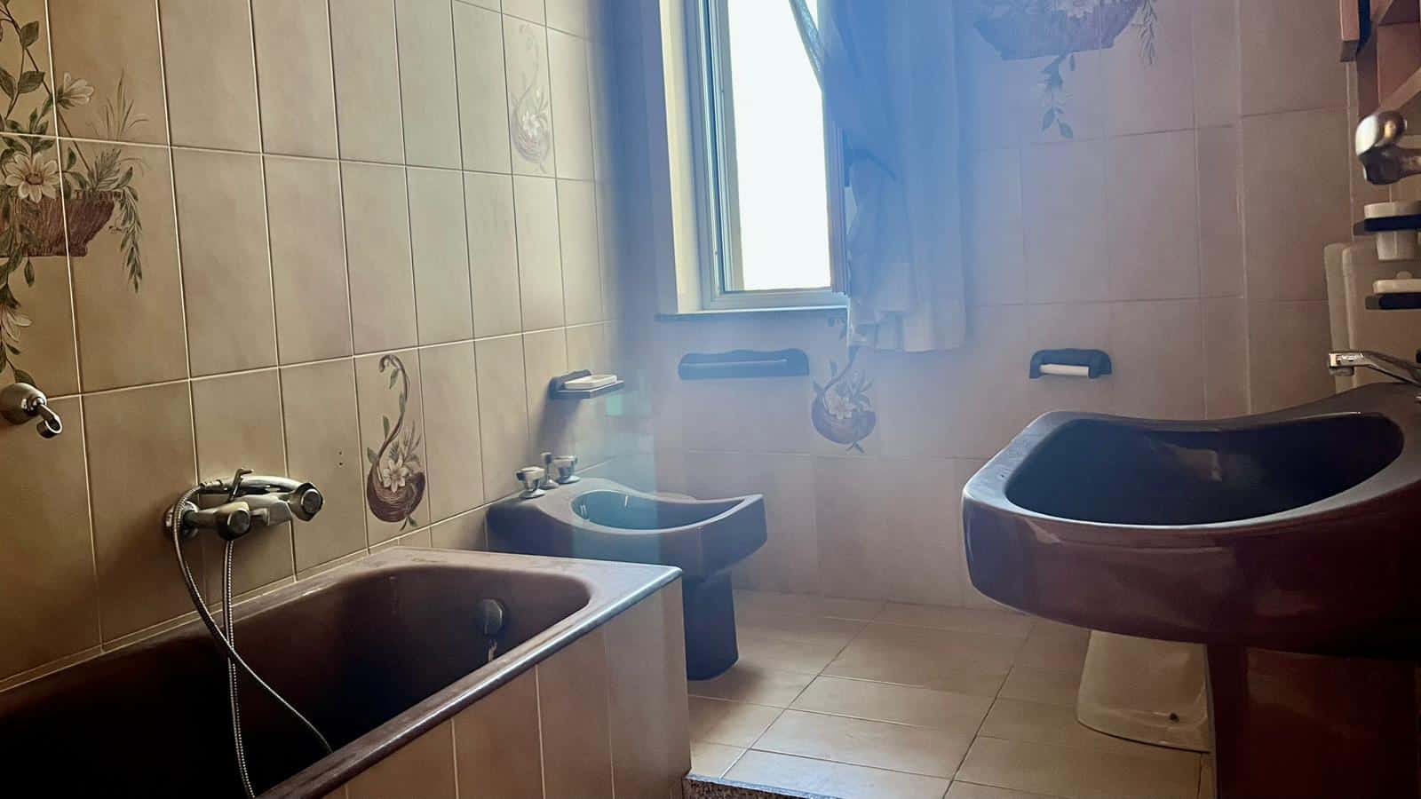 Lavabo e bidet in bagno con pareti rivestite di ceramica decorata e finestra che lascia entrare luce naturale. Spazio funzionale con accessori bagno e vasca.