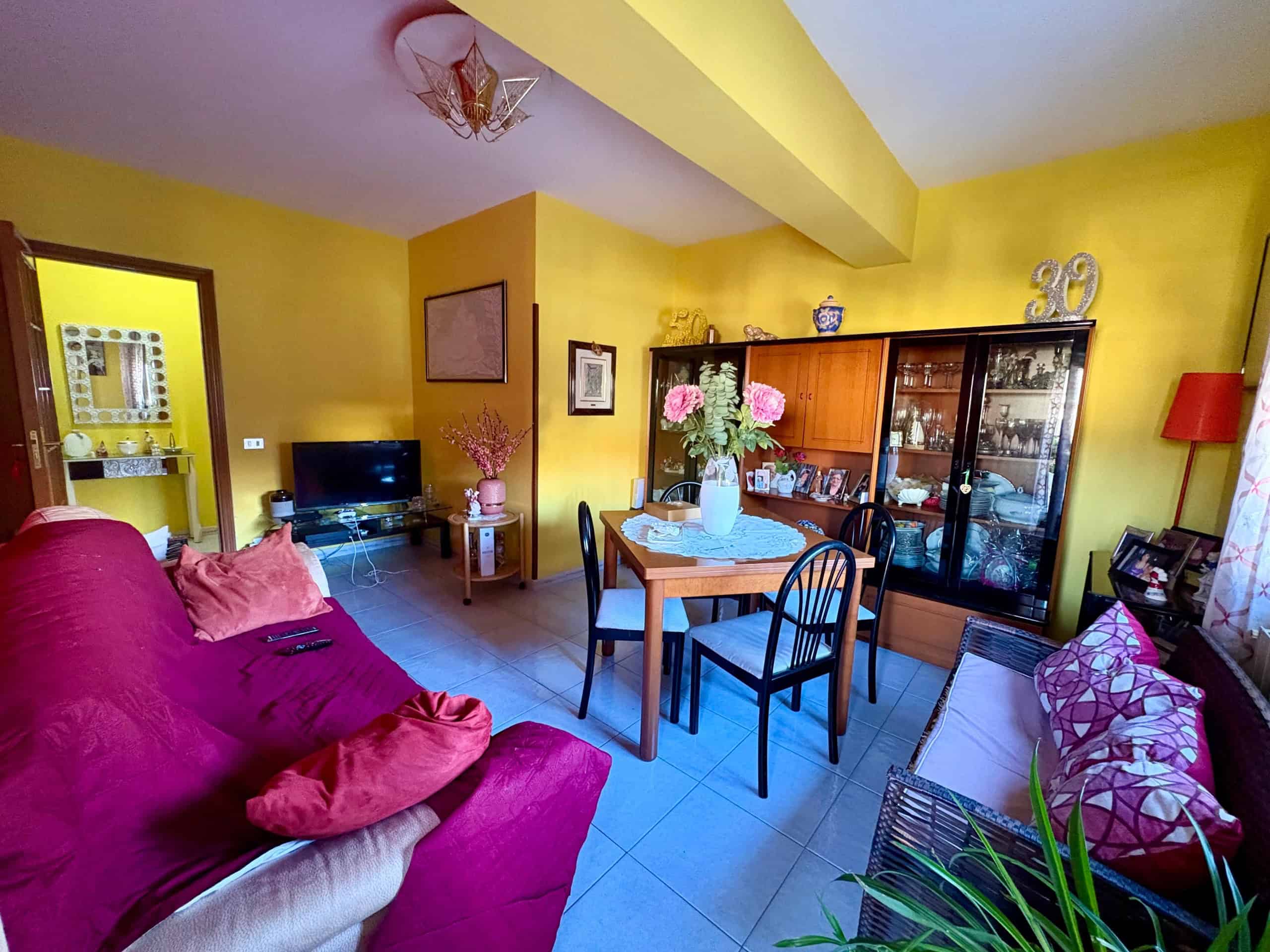 Divano rosso in soggiorno con pareti gialle e mobili in legno, tavolo con fiori e accessori decorativi, ambiente accogliente e luminoso, ideale per la casa e l’arredamento.