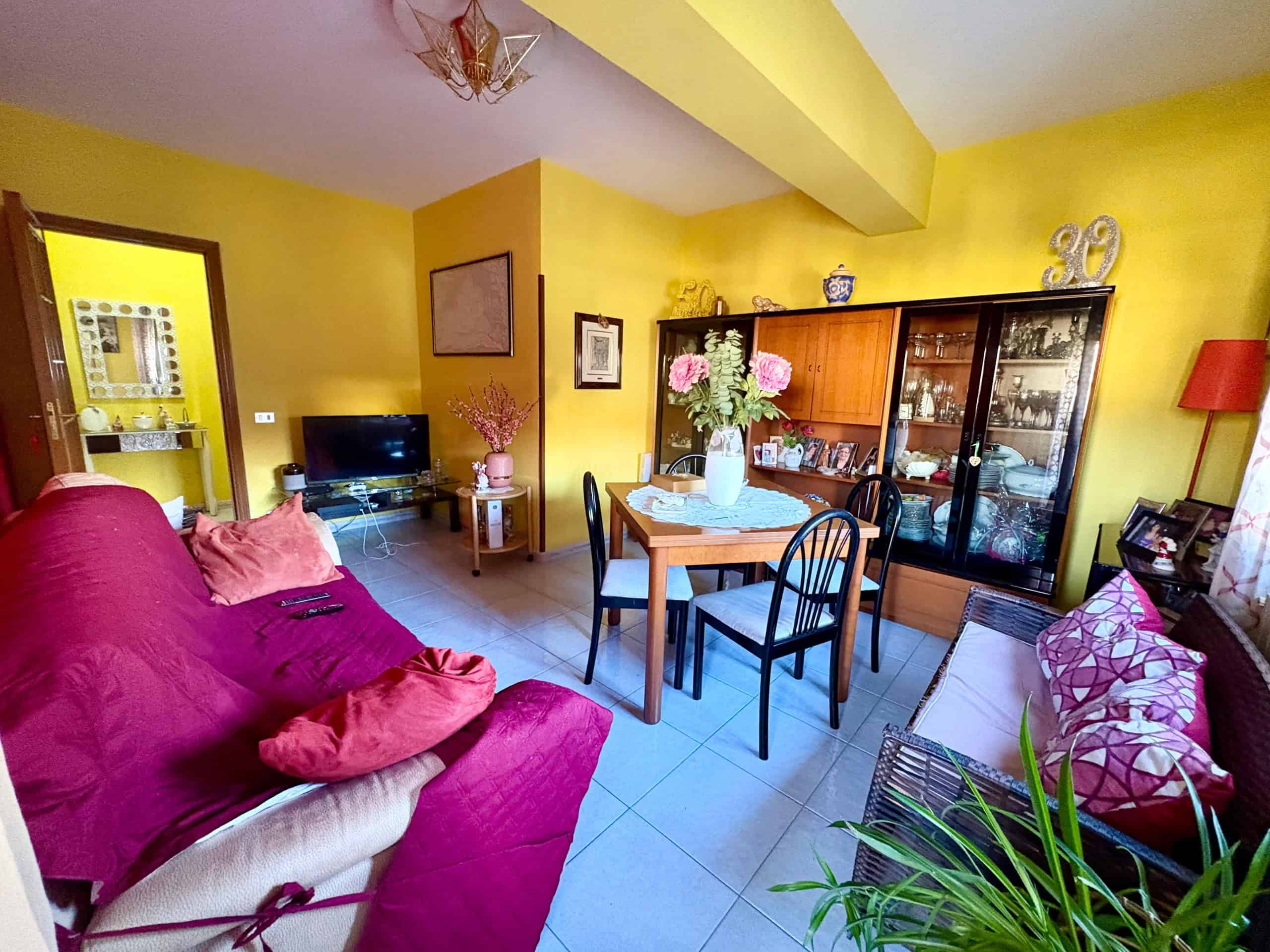 Accogliente soggiorno con arredi vintage, divani colorati e mobili in legno, illuminato da luce naturale e decorato con piante e oggetti d'antiquariato, ideale per vivere momenti di relax e convivialità.