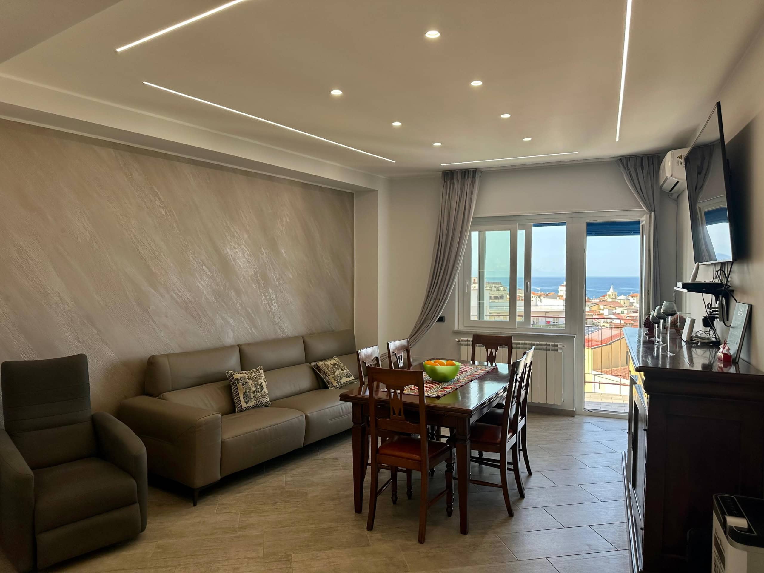Vista di soggiorno elegante con parete decorativa, ampio divano, e vista mare dall'ampia finestra, atmosfera luminosa e moderna, ideale per case di lusso a NextCasa.