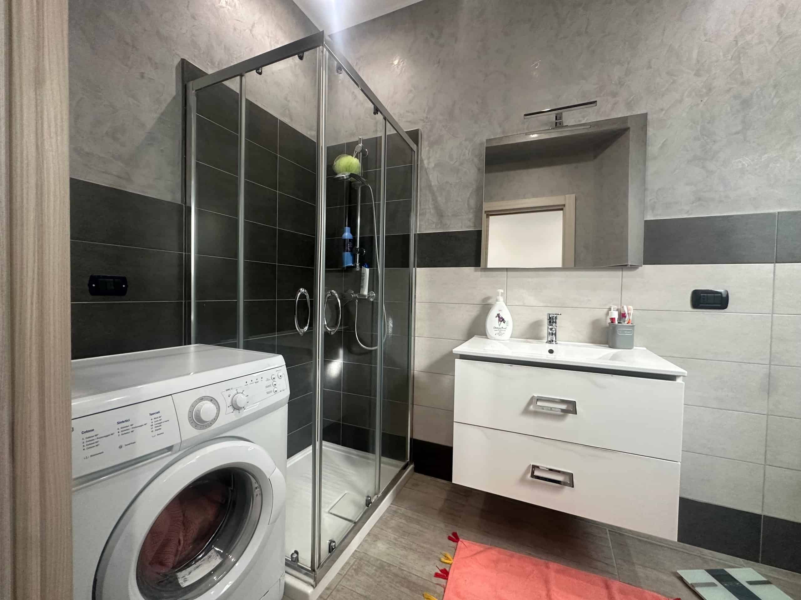 Lavanderia moderna con cabina doccia, lavabo bianco con specchio, phon e prodotti per l'igiene, in ambienti funzionali e dal design elegante, ideale per case in vendita o affitto.