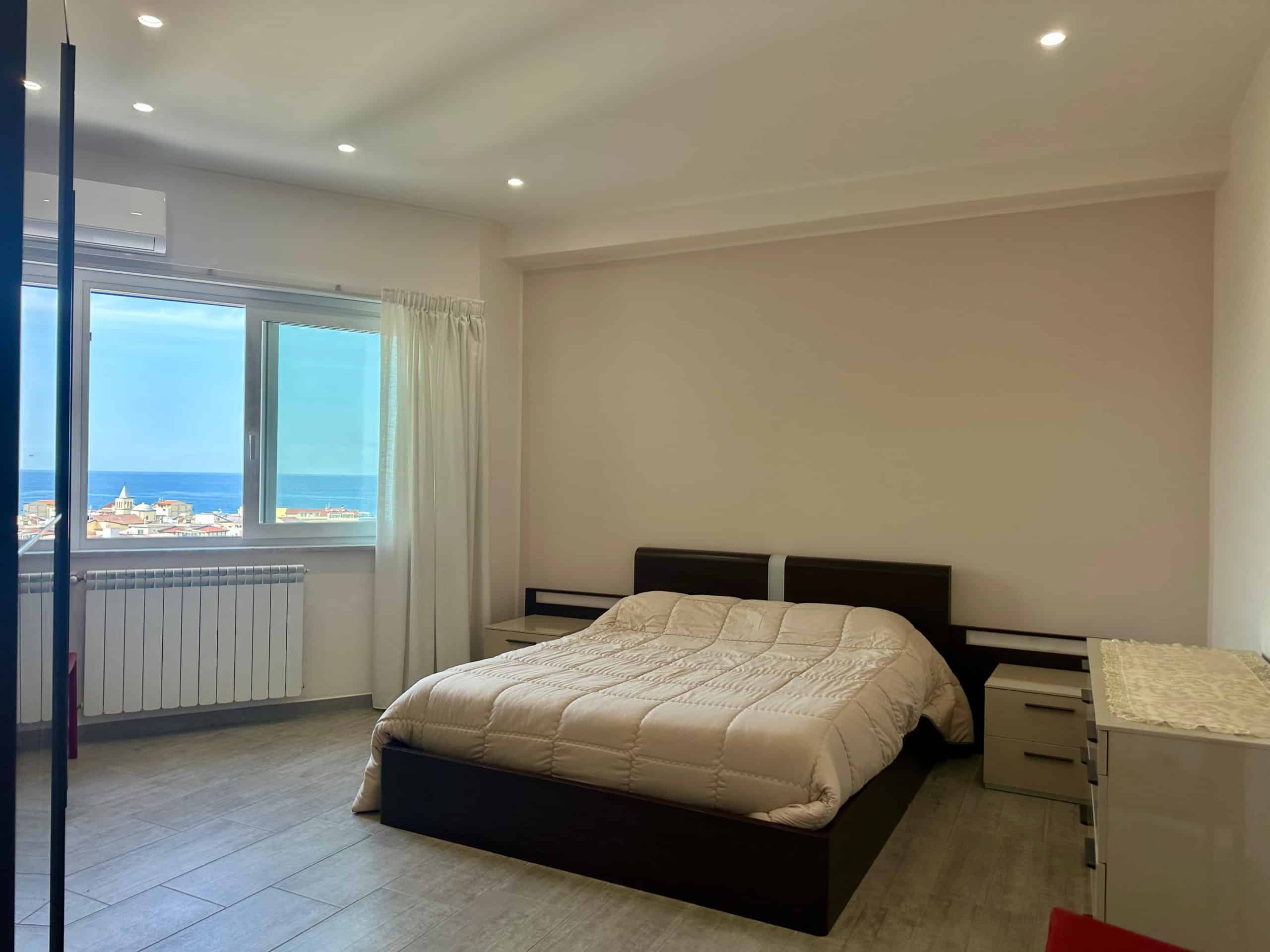 Vista di una camera da letto moderna con vista mare, grande finestra con tende chiare, pareti bianche, letto matrimoniale con coperta beige, mobili in stile minimalista, ideale per immobili in vendita o affitto.