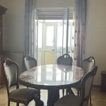 Mobili soggiorno classici con tavolo in marmo e sedie imbottite, armadio anticato in legno scuro, illuminazione naturale da porte finestre con tendaggi decorativi, stile tradizionale e atmosfera accogliente.