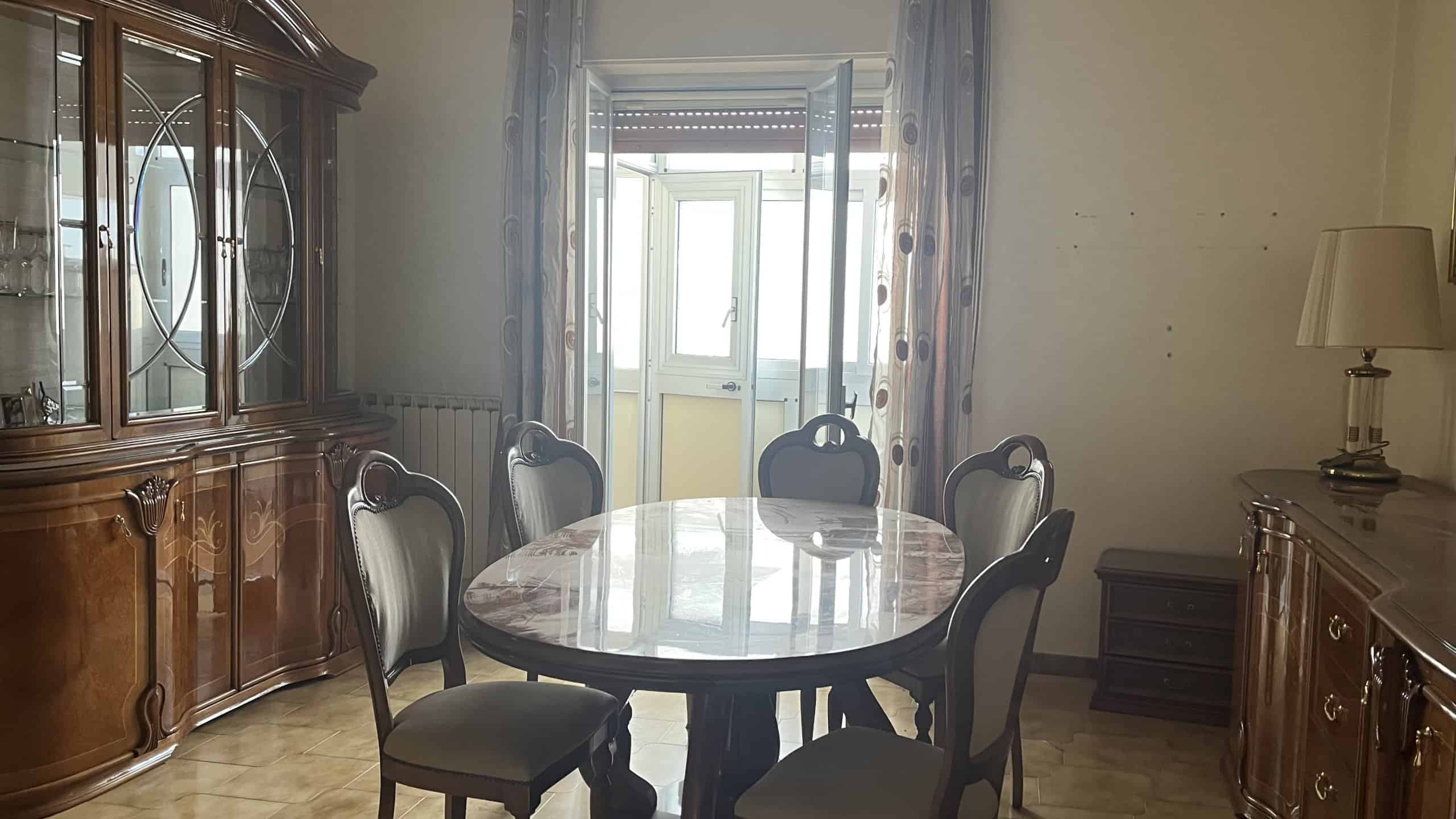 Mobili soggiorno classici con tavolo in marmo e sedie imbottite, armadio anticato in legno scuro, illuminazione naturale da porte finestre con tendaggi decorativi, stile tradizionale e atmosfera accogliente.