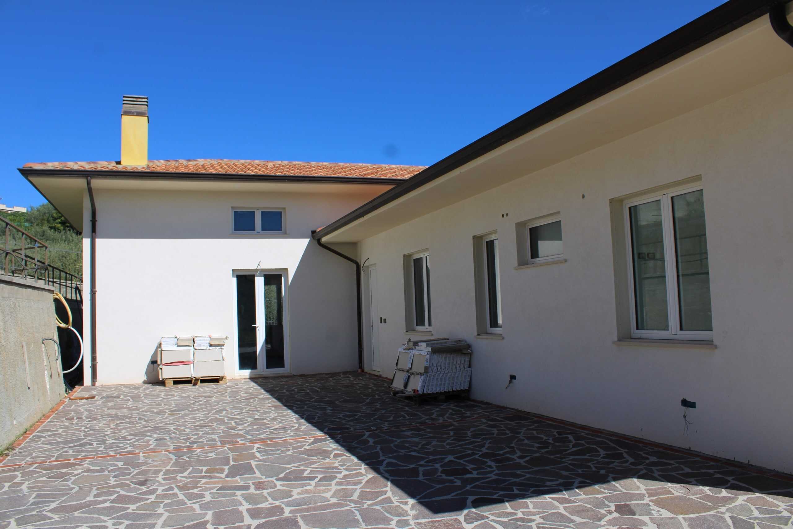 F）。vista esterna di una casa moderna con cortile in pietra, pareti bianche, finestre e porta scorrevole, cielo sereno, progetti di ristrutturazione in corso, soluzione abitativa in fase di completamento, NextCasa.
