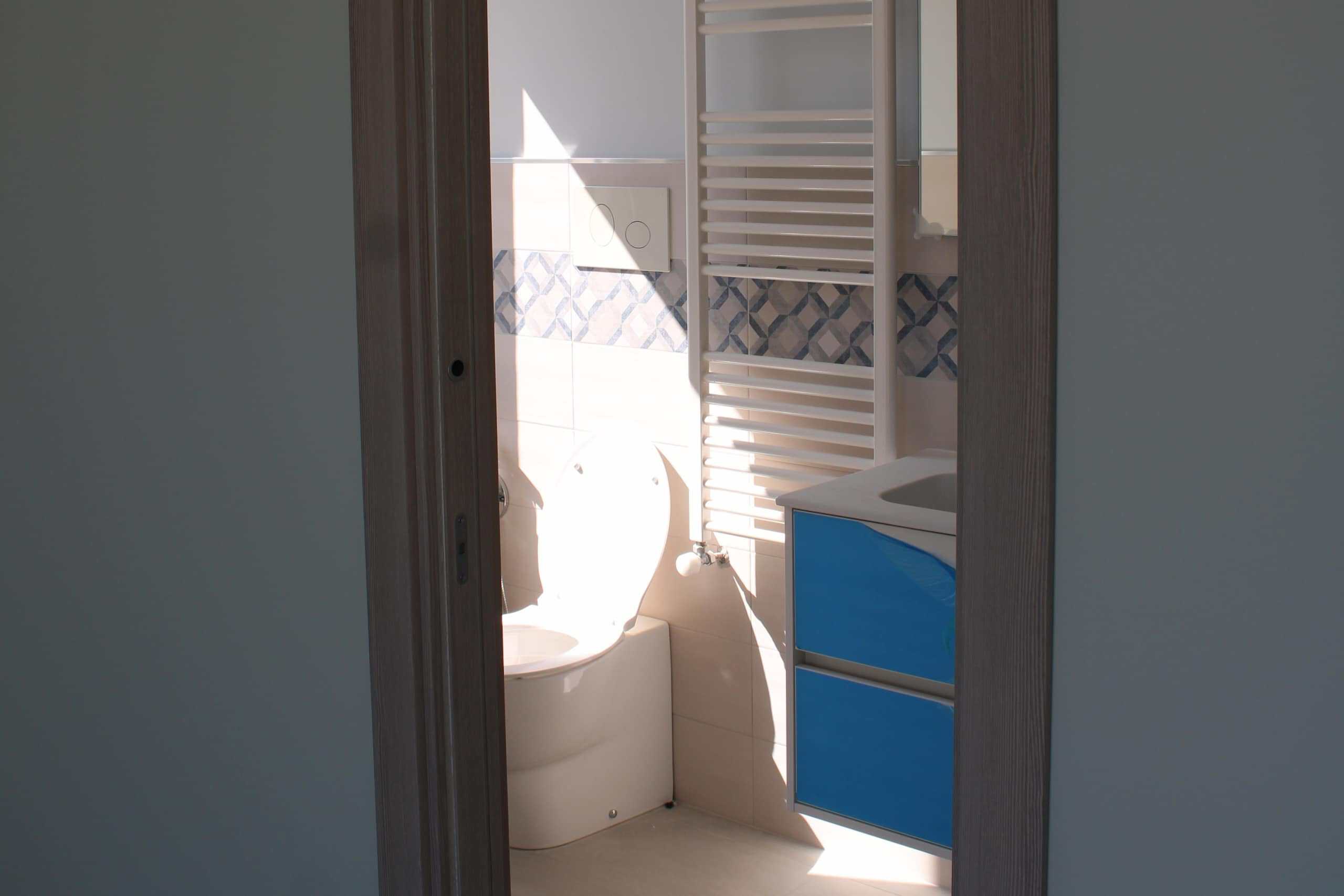 WC bagno privato con piastrelle moderne, lavabo blu e scaldasalviette bianco, vista attraverso porta in legno, ambiente luminoso e funzionale.