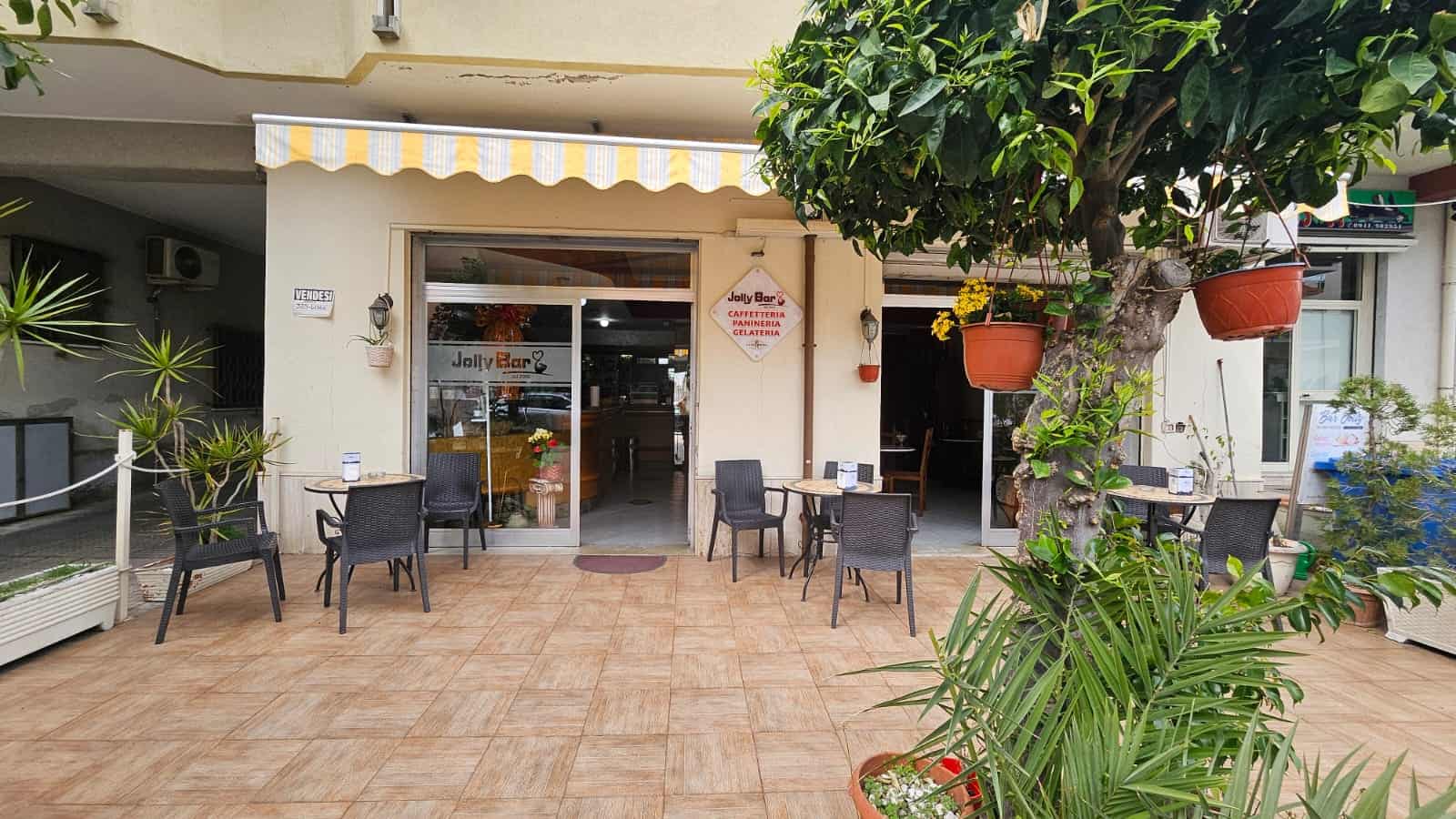 Piazza esterna di caffetteria con tavolini e sedie, ambiente accogliente con piante e decorazioni, ideale per rilassarsi e gustare un gelato o una bevanda all'aperto.