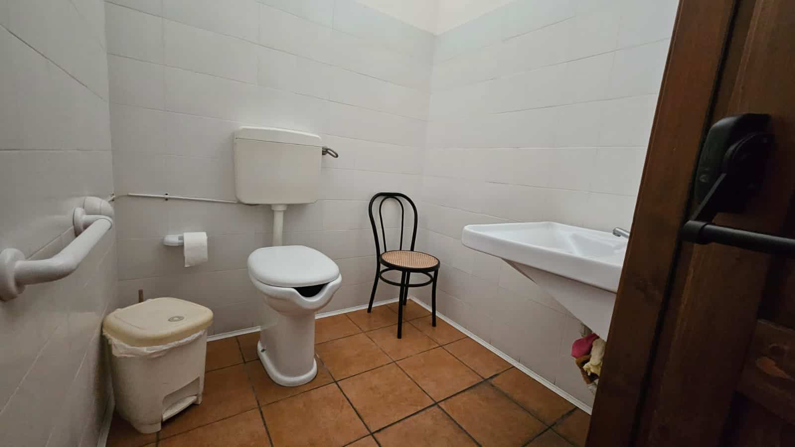 Bagno accessibile con WC e lavabo in stile semplice, arredato con sedie e accessori pratici, ideale per esigenze di comfort e funzionalità in abitazioni o strutture ricettive.