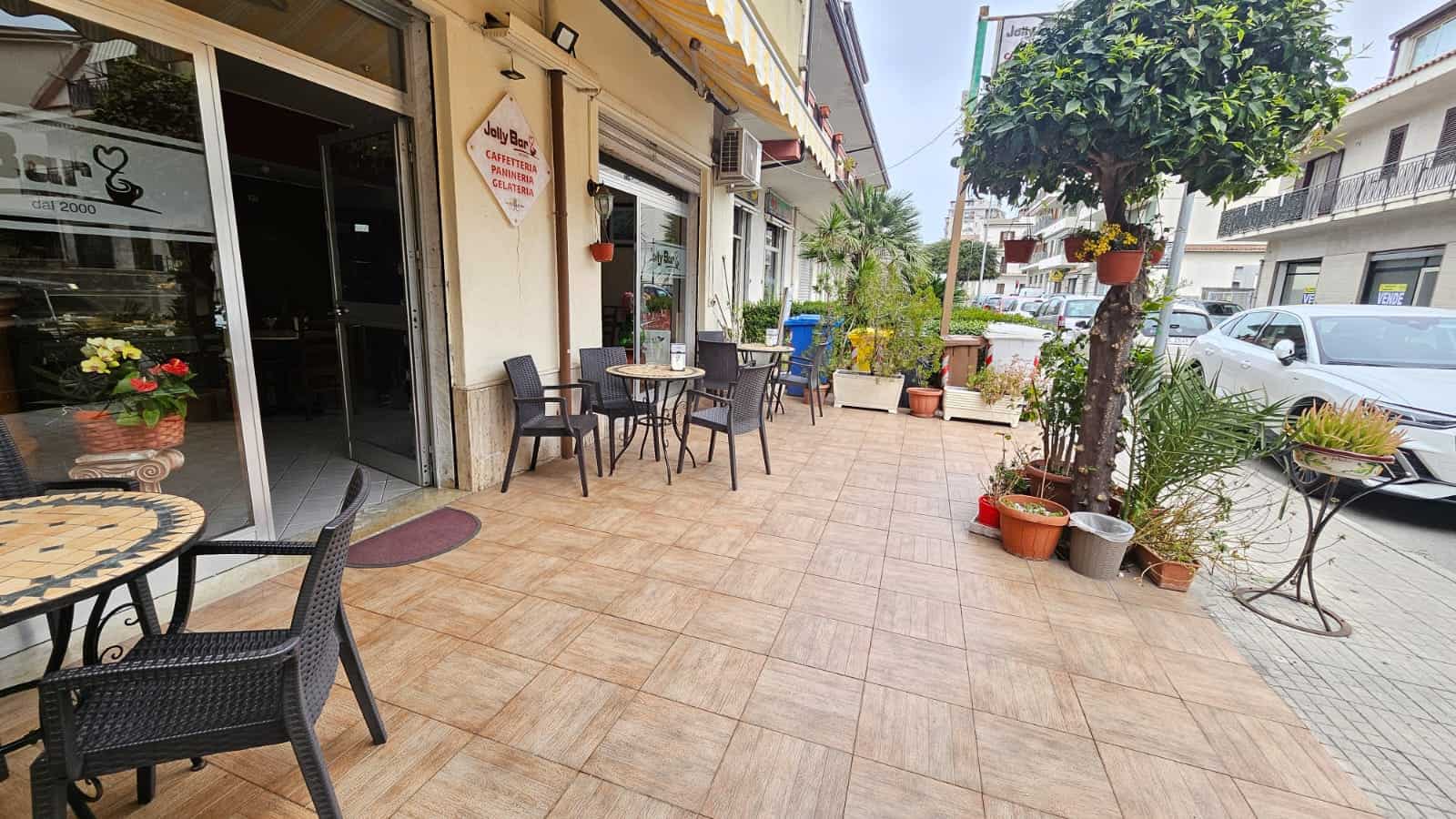 Vassoio di tavoli e sedie in esterno davanti a caffetteria con piante e arredi rustici, atmosfera accogliente per colazioni e pranzi all'aperto.