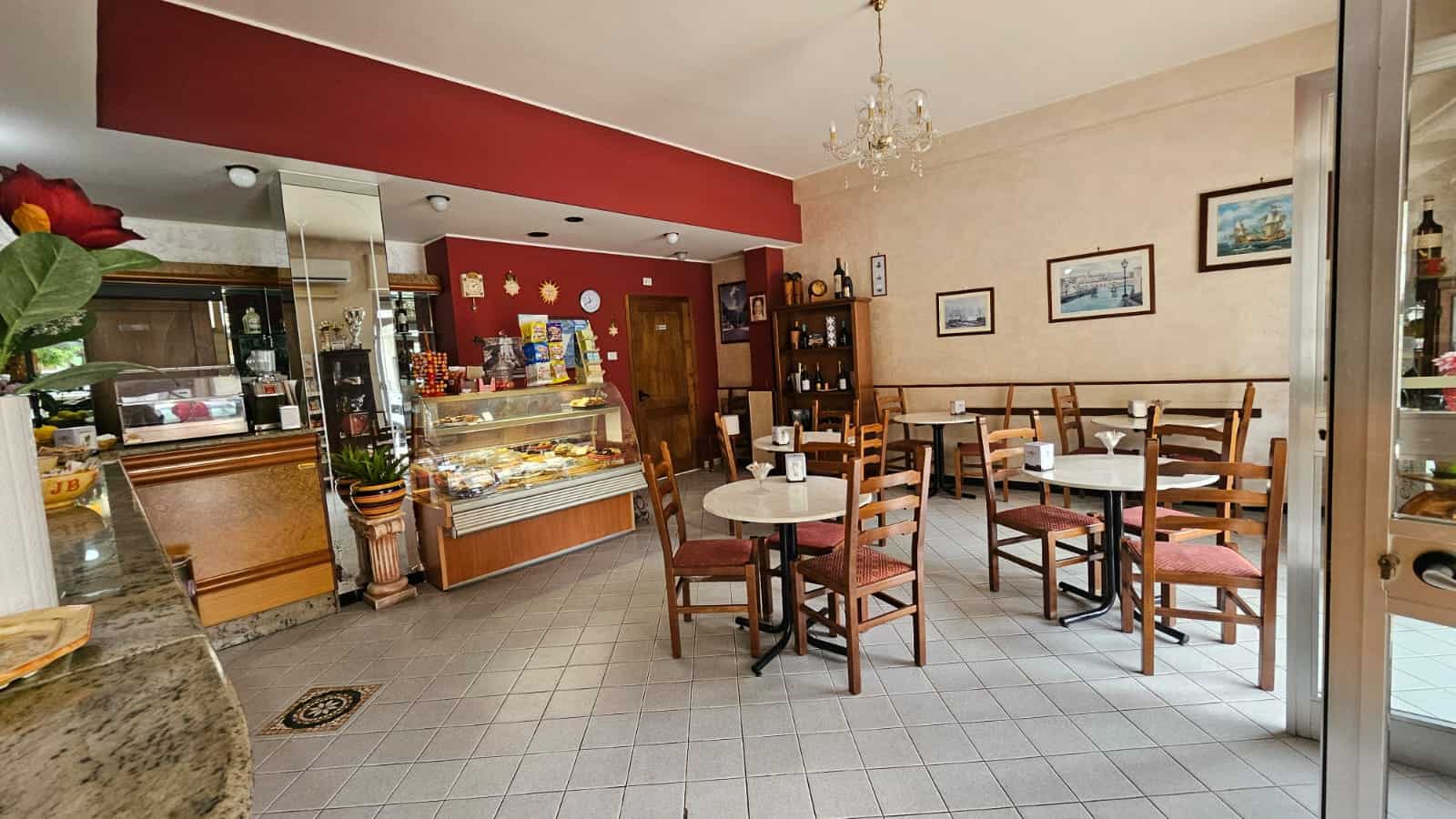 Sedie e tavoli in un accogliente caffetteria con pareti decorate e esposizione di dolci e snack in vetrina, ideale per momenti di relax e colazioni.