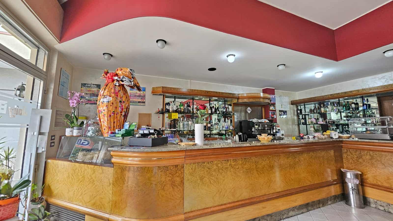 Banco bar in un ristorante con struttura in legno, scaffalature con bottiglie di vino e alcolici, decorazioni floreali e ambiente accogliente, ideale per attività di ospitalità e ristorazione.