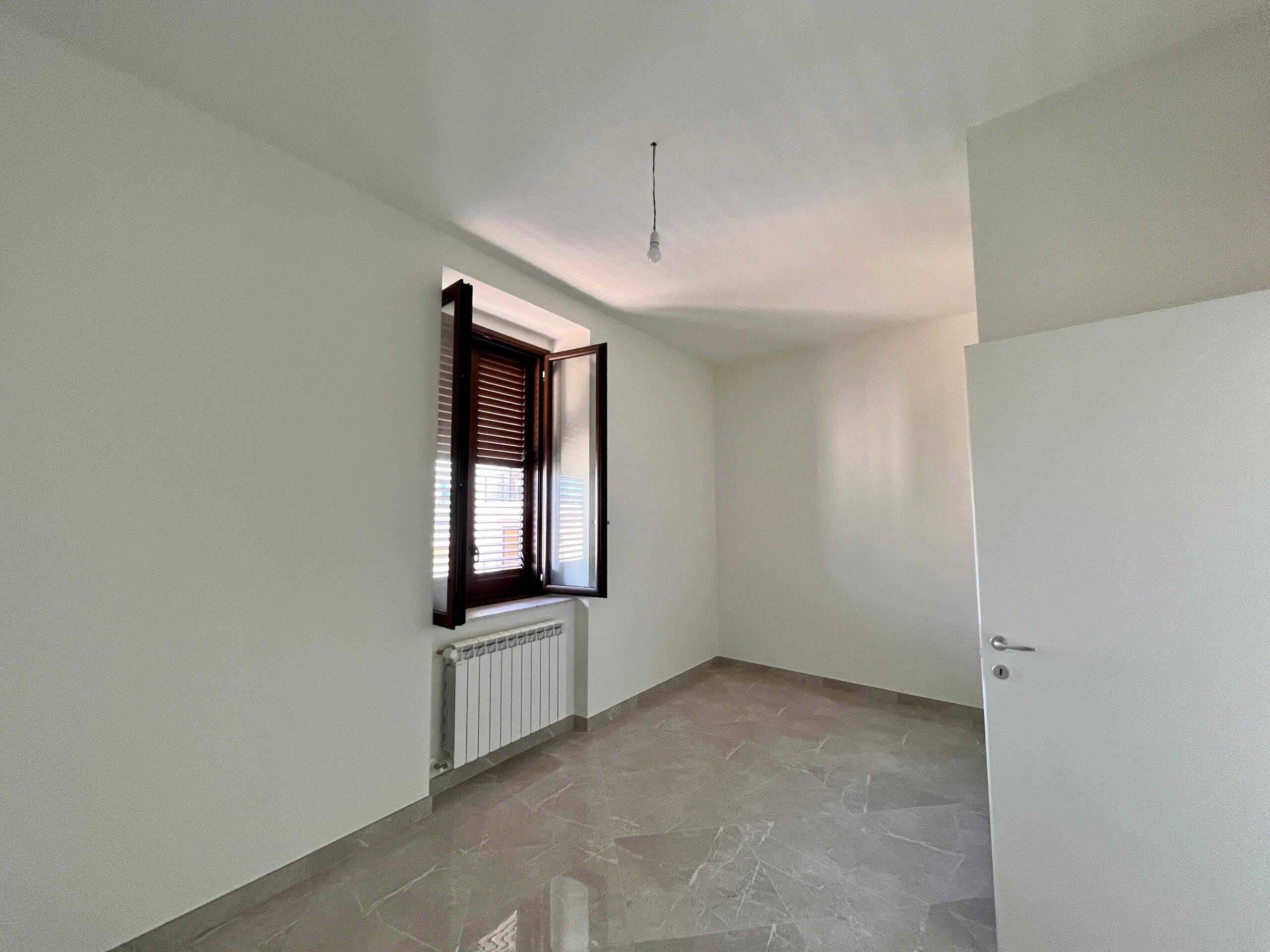 Finestra con tapparelle in legno in una stanza vuota, pareti bianche e pavimento in gres chiaro, ambienti moderni, ideale per ristrutturare o creare una casa personalizzata.