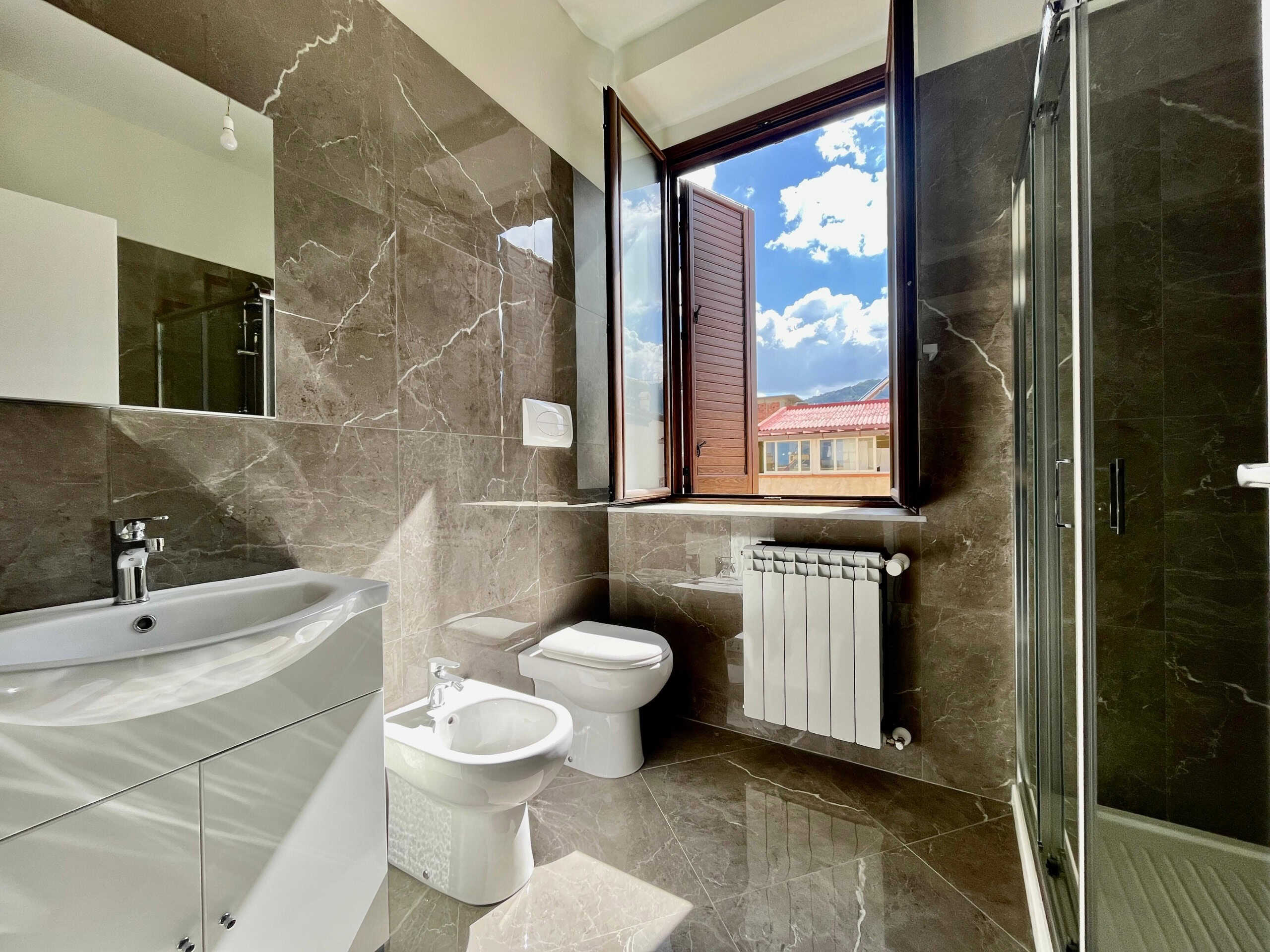 Lavabo e servizi in bagno con pareti e pavimenti in marno, finestra aperta su vista panoramica delle case e cielo. Illuminazione naturale, ambiente moderno e raffinato.