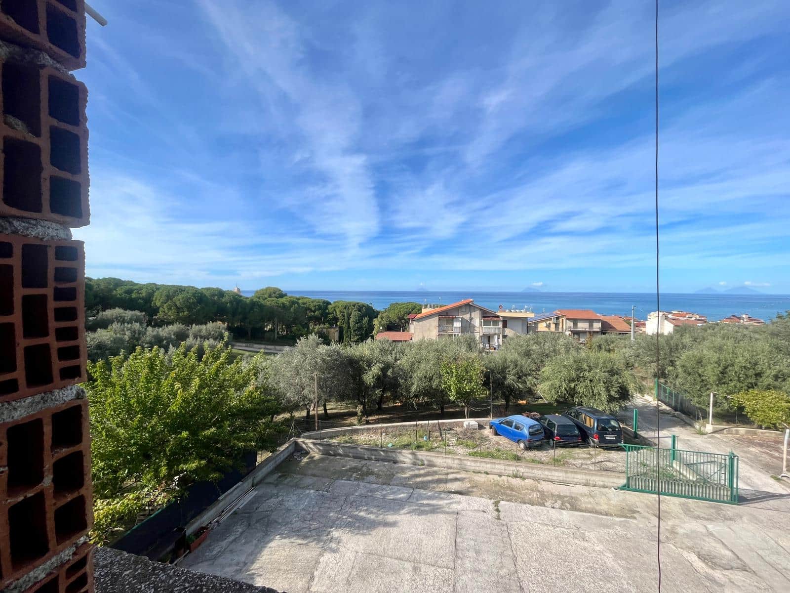 Vista mare e paesaggio residenziale dal balcone di un appartamento NextCasa, con cielo azzurro e nuvole leggere, durante una giornata soleggiata in una località costiera italiana.