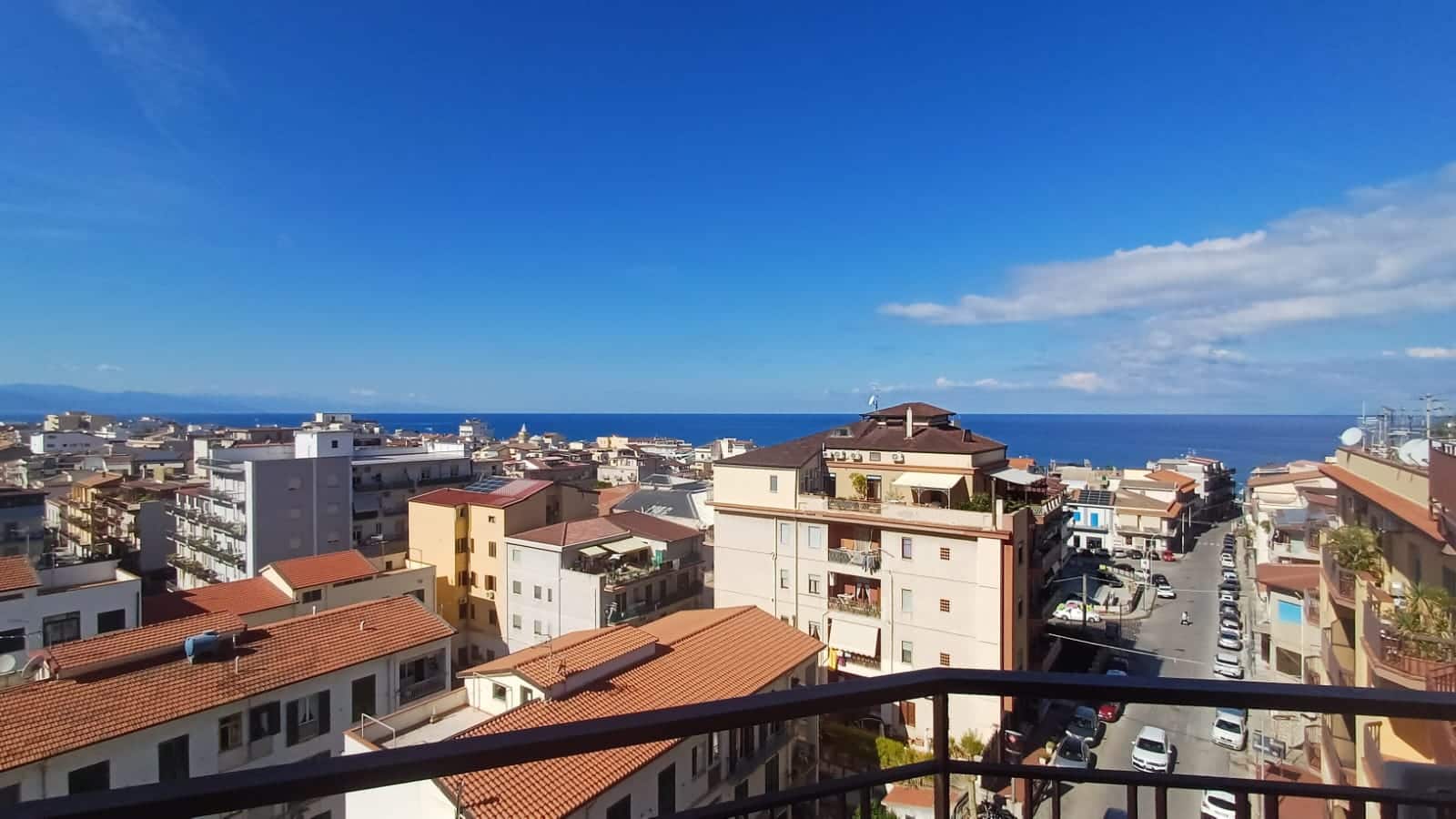 Vista mare e skyline delle abitazioni nel centro di una città costiera, vista da un balcone con ringhiera in ferro, con edifici moderni e tradizionali, cielo sereno e mare blu brillante.