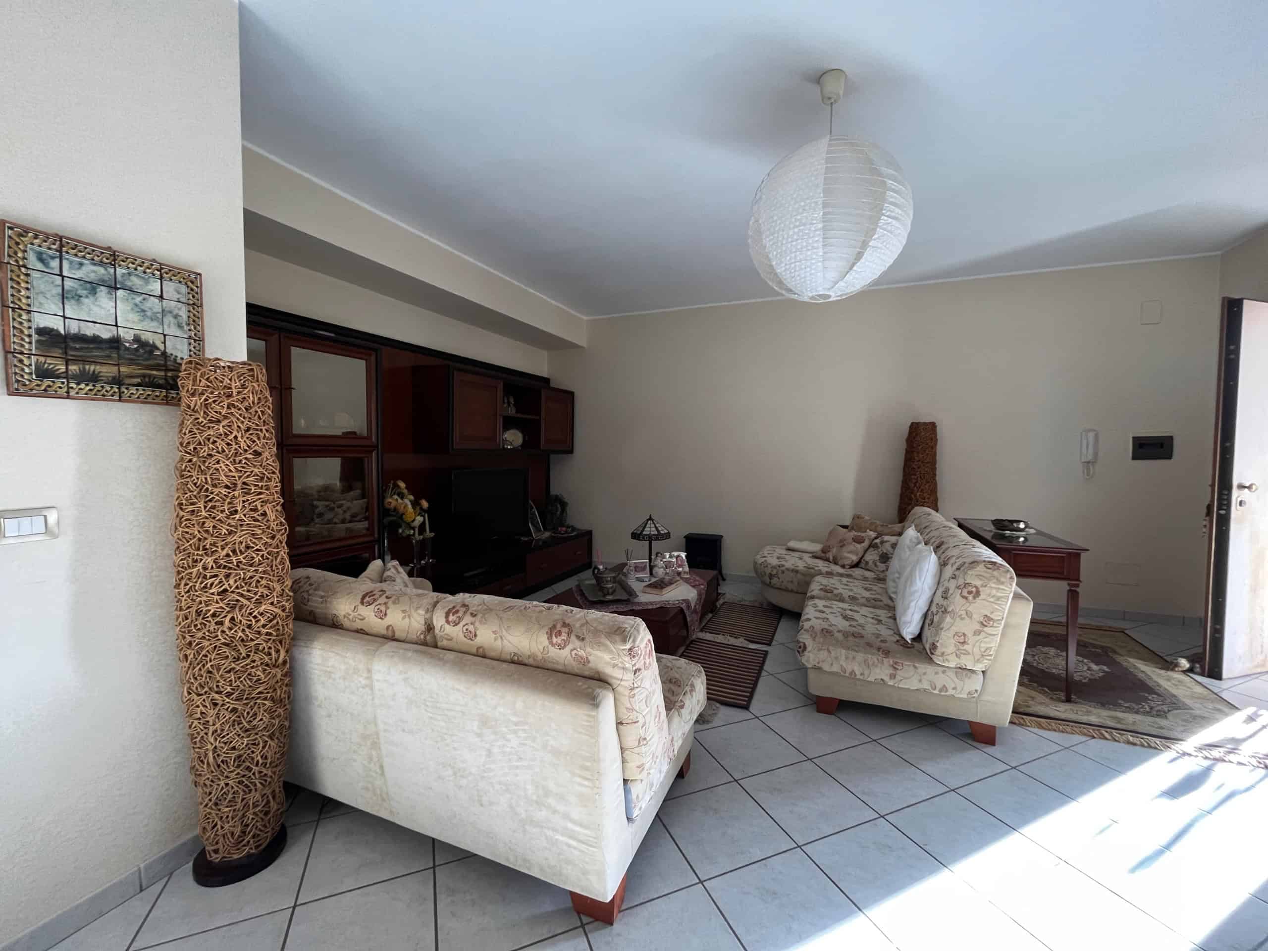 Accogliente soggiorno con divani in tessuto fiorato, mobile parete in legno scuro e oggetti decorativi, illuminato da luce naturale e lampada a sospensione, ideale per vivere momenti di relax.