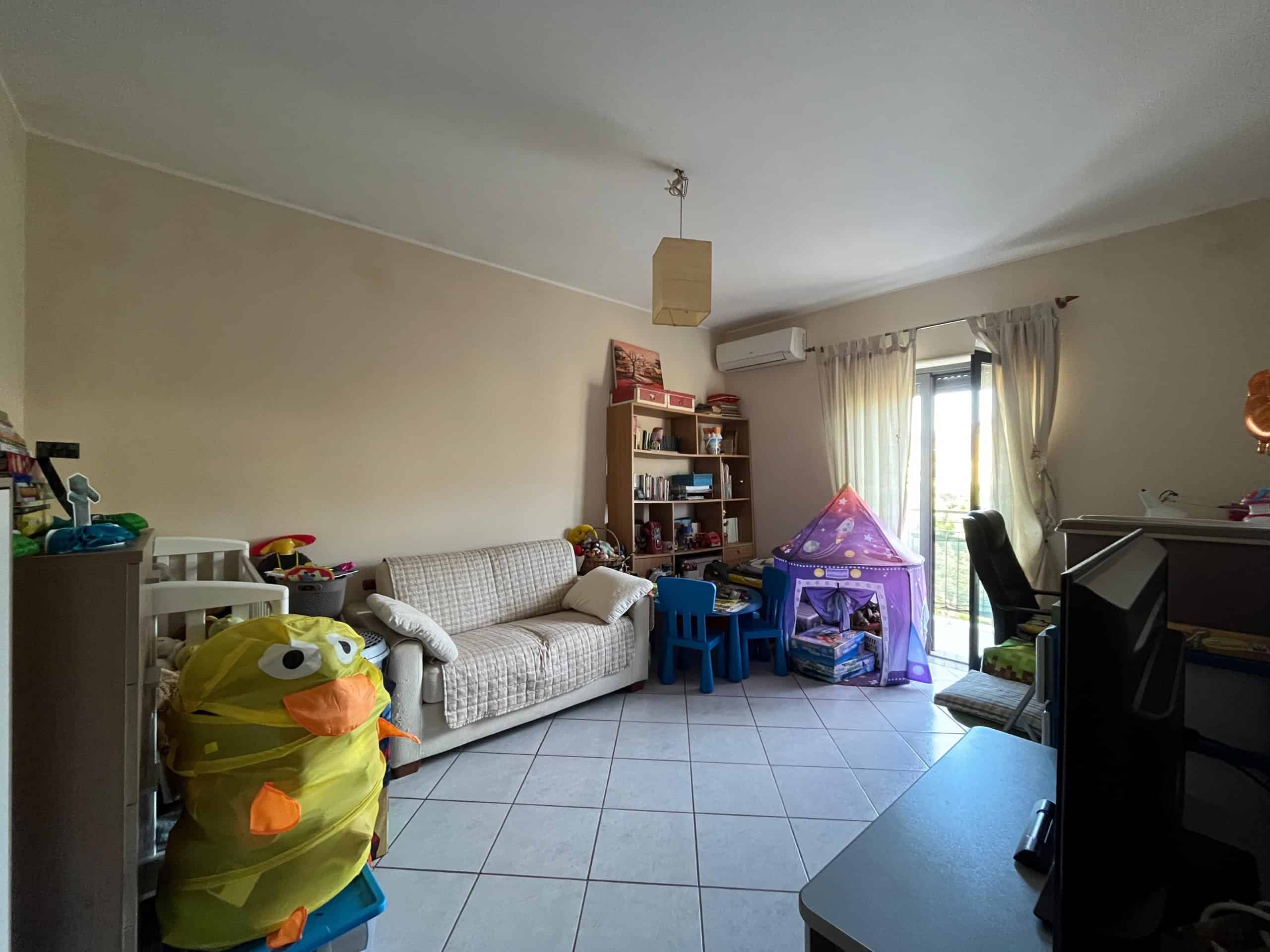 Ampia stanza soggiorno luminosa con giochi e arredi per bambini, divano, scaffale, tenda e accessori per giochi nella casa NextCasa. Perfetta soluzione di arredamento per famiglie.