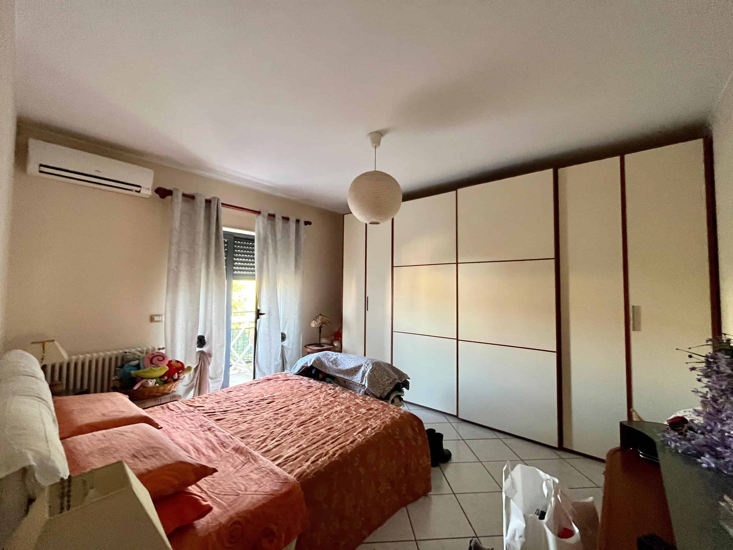 Accogliente camera da letto con arredamento moderno, grande armadio a muro e balcone con vista. Atmosfera luminosa e calda, ideale per un abitativo confortevole e funzionale.