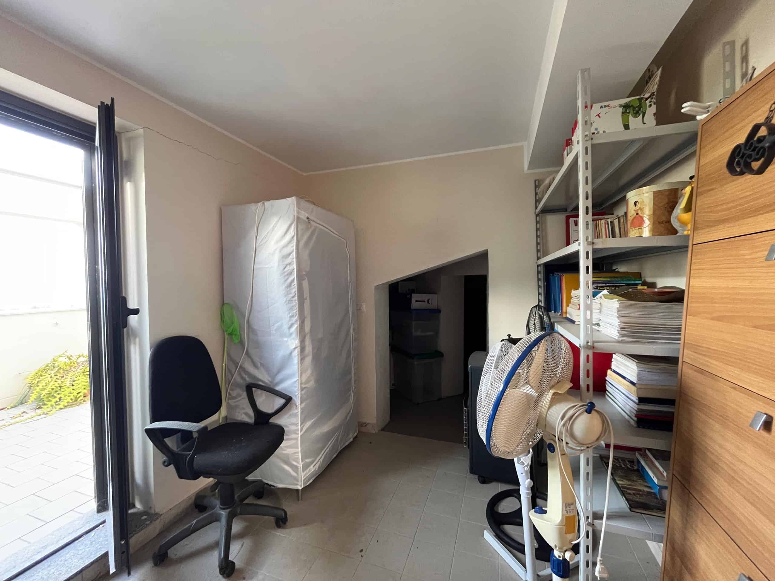 Immagine di una stanza di casa trasformata in ufficio o studio con scaffali pieni di documenti e libri, sedia da ufficio, ventilatore e accessori vari, ideale per smart working o studio casalingo.