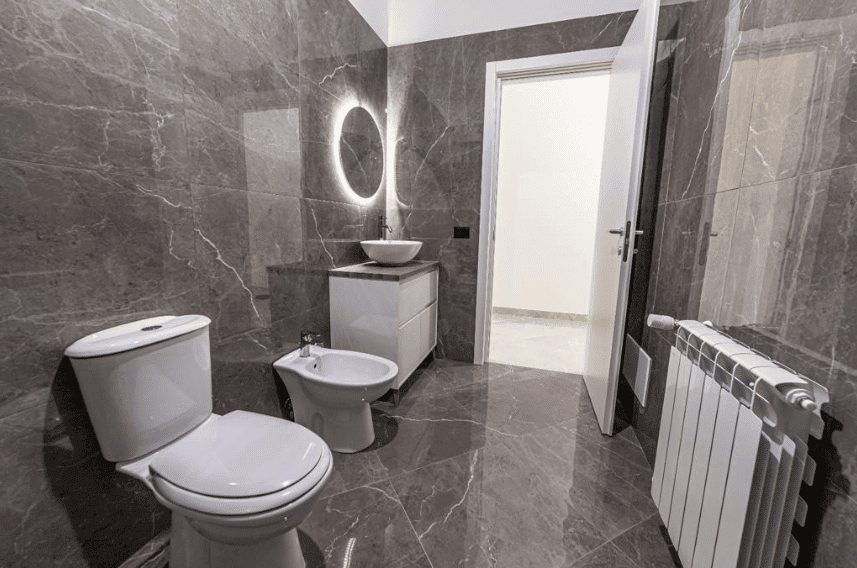 Bagno moderno con pareti e pavimenti in marmo grigio, lavabo con luce a LED, sanitari bianchi, arredo semplice e funzionale, porta aperta che porta a un'altra stanza, riscaldamento a radiatore in acciaio inox.
