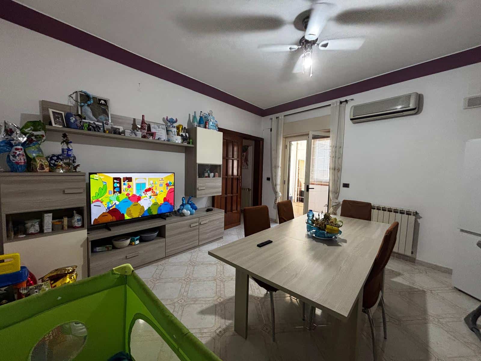 Interno soggiorno con tavolo, sedie e mobili, decorazioni e TV, ambiente luminoso e accogliente.