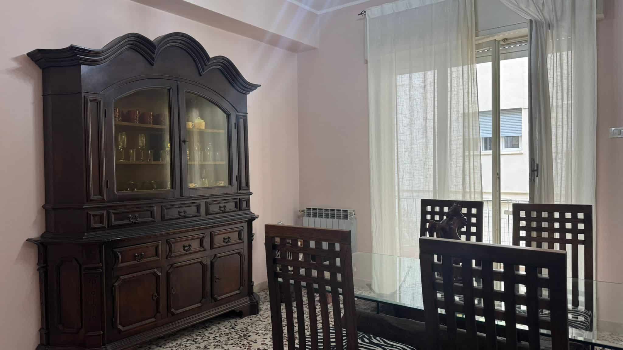Antico mobile in legno scuro in soggiorno con vetrina e mobile basso, vicino a grandi finestre con tende leggere, ambiente luminoso e accogliente con dettagli di design classico.
