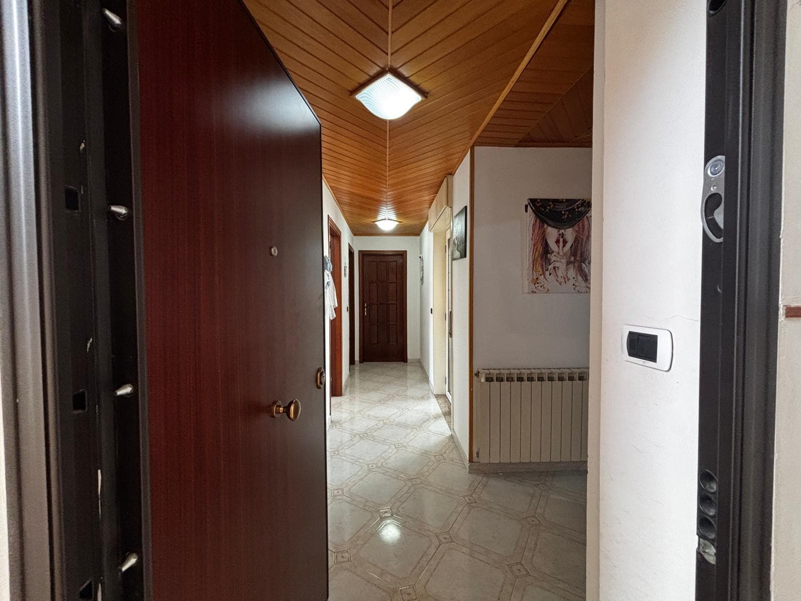 Ingresso di casa con pavimento in marmo, pareti bianche, soffitto in legno, illuminazione led, arte moderna e radiatore; imponente porta d'ingresso in legno con maniglia antica, camera accogliente e funzionale.