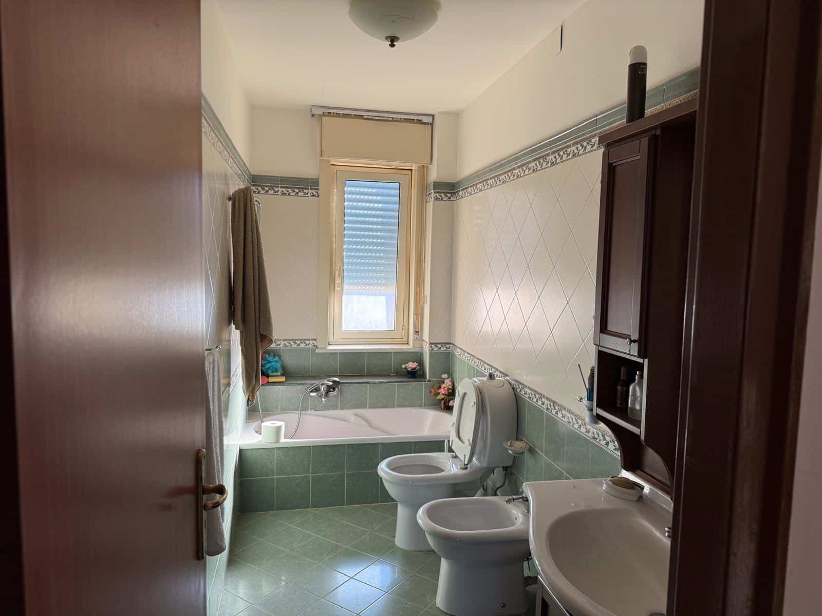 Ampia stanza da bagno con vasca, bidet, WC e lavabo, finestra per illuminazione naturale, rivestimenti in piastrelle bianco e verde, dettagli decorativi, arredi in legno scuro, ambiente ben tenuto e funzionale.