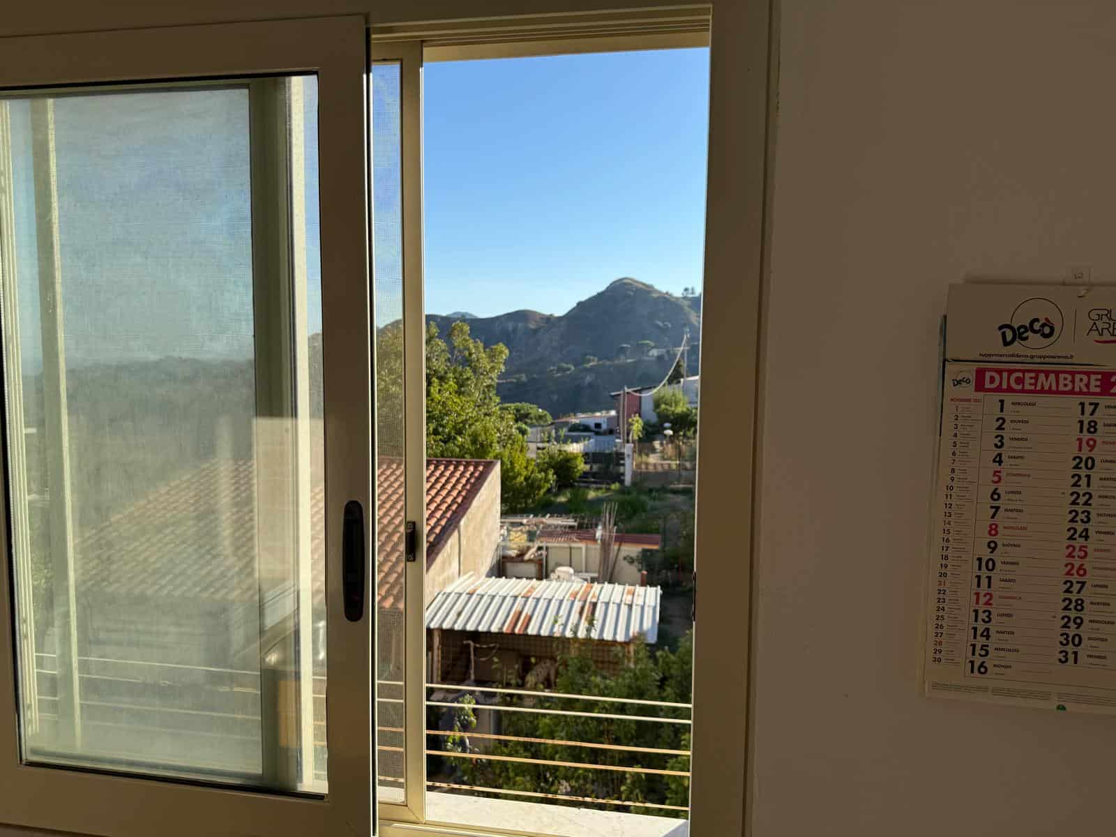Finestra aperta con vista panoramica su montagne e natura, illuminazione naturale in un ambiente abitativo, vista su colline e cielo sereno, scatto interno di un appartamento in vendita o affitto.